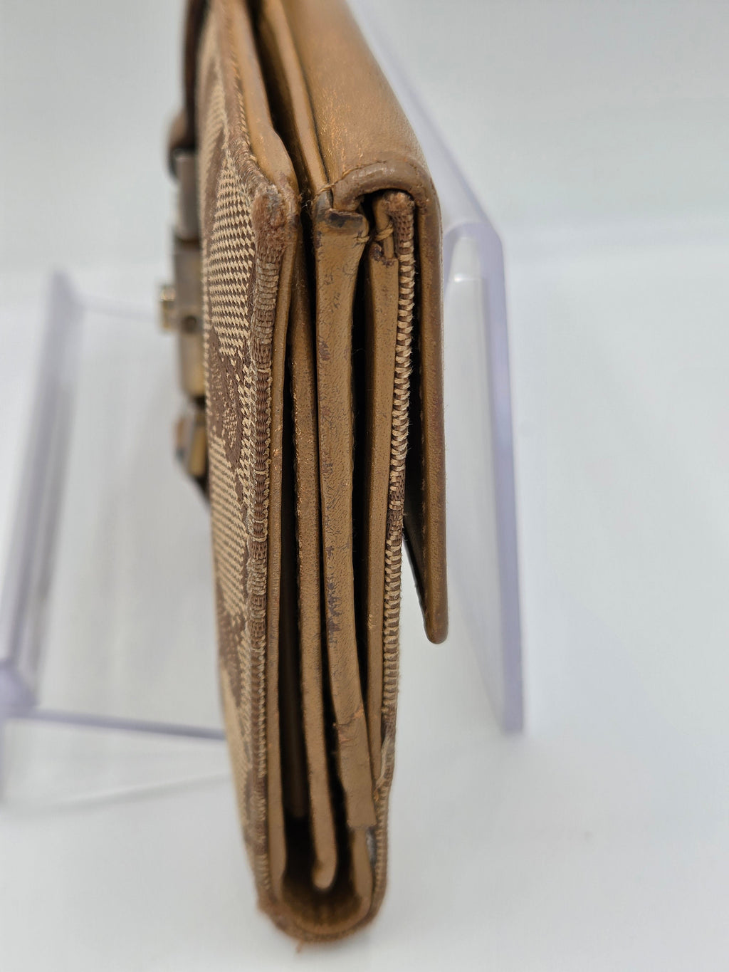 A close-up side view of a tan leather wallet with a textured woven pattern, resting on a clear acrylic display stand against a white background.