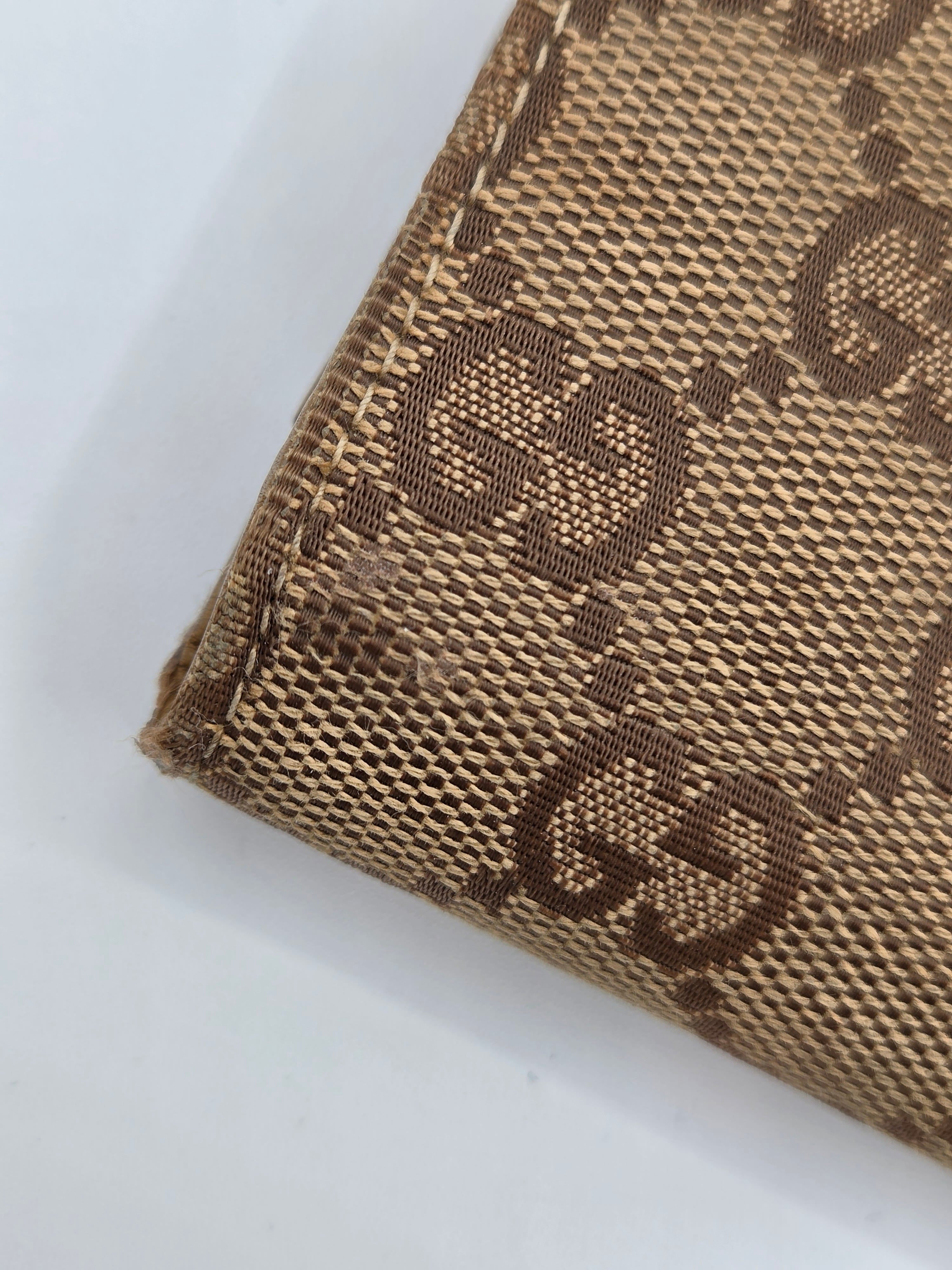 Close-up of a beige and brown monogram canvas material with a woven pattern, showing part of a folded item on a white background.