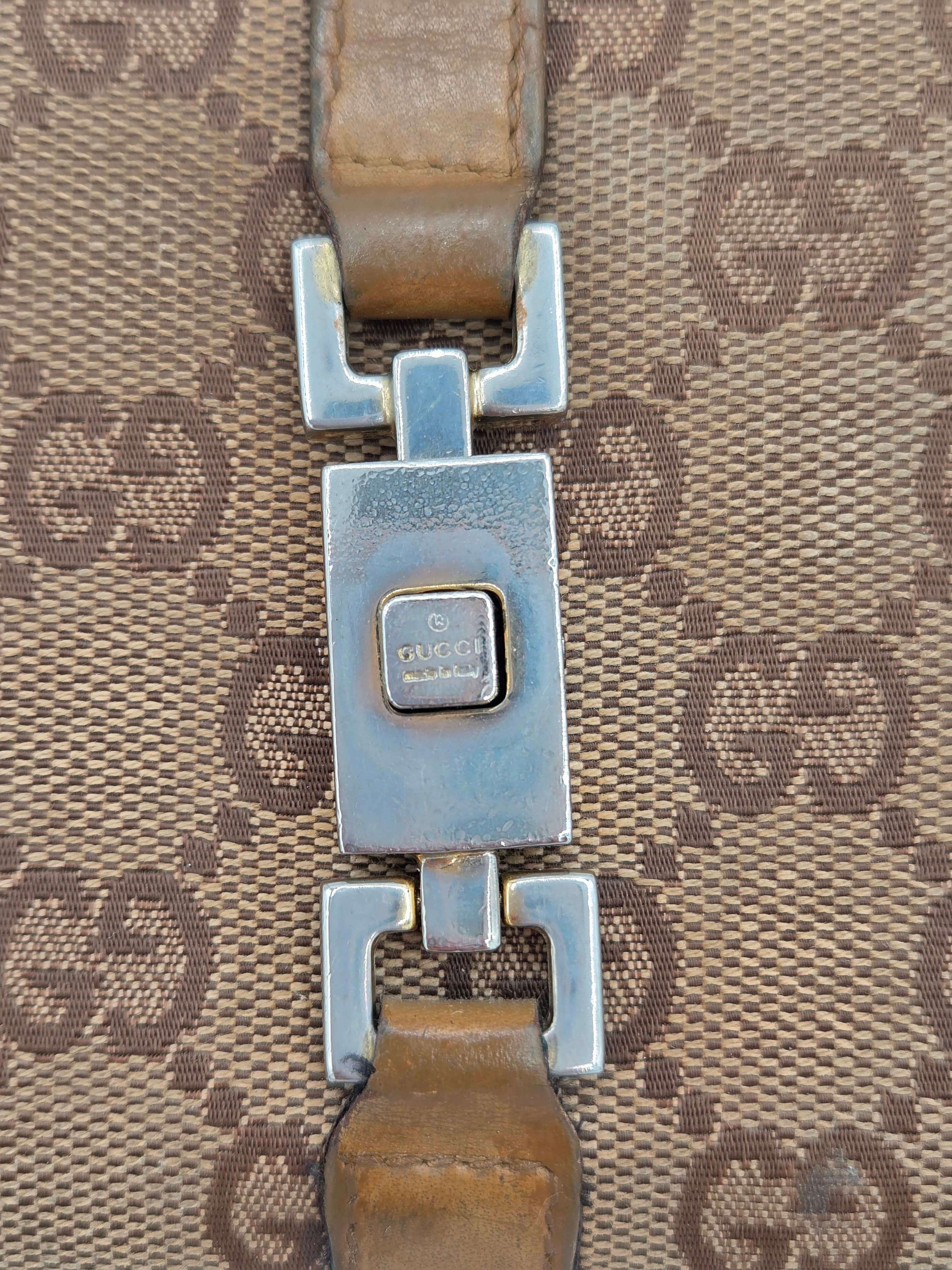 Close-up of a Gucci monogram bag strap with a silver-tone metal clasp and brown leather on a beige GG canvas background.
