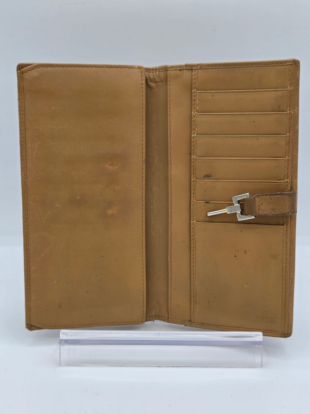 A tan leather wallet shown open, featuring multiple card slots, a central keyhole pocket with a silver key, and a large slip pocket below. A clear plastic sleeve is positioned to the right of the wallet on a white background.