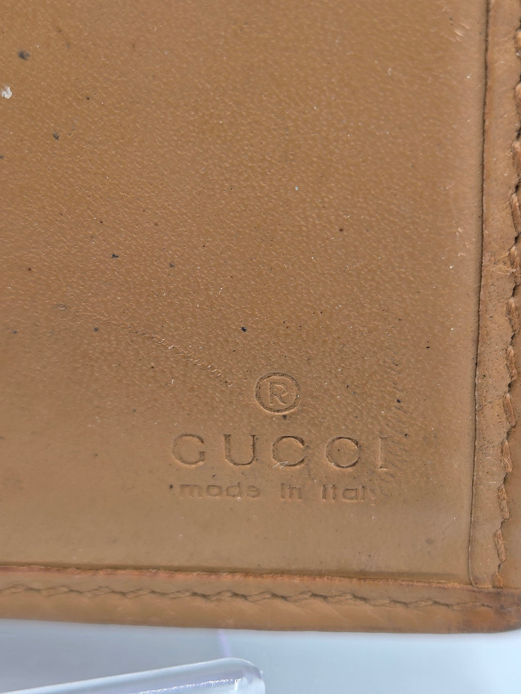 Close-up of a tan leather wallet corner with visible Gucci branding embossed on the surface and stitched edges.
