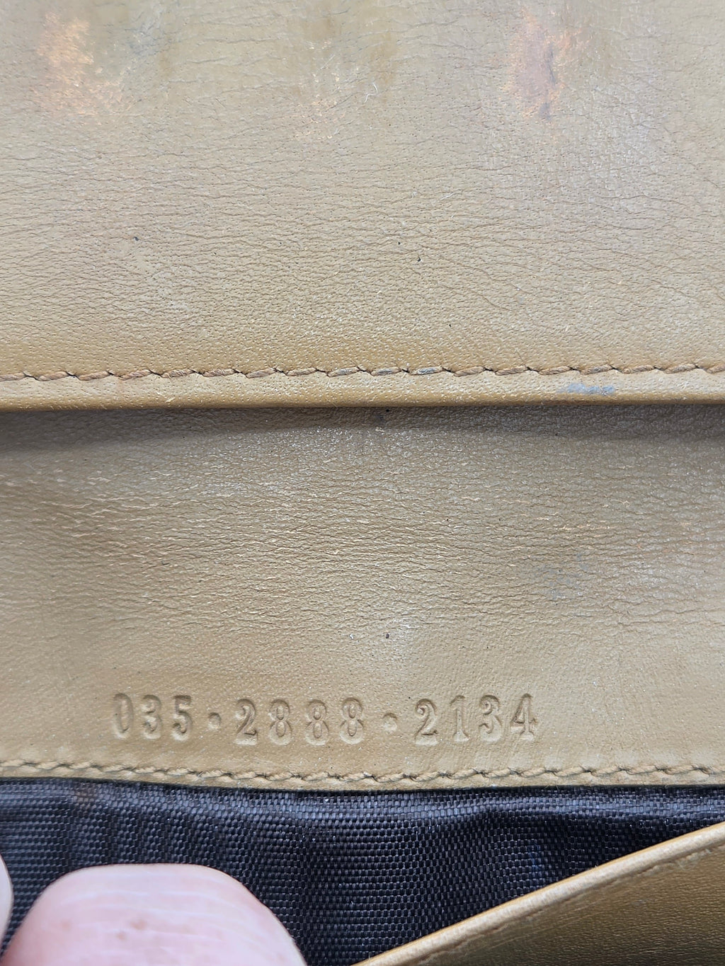 Close-up of a tan leather wallet or clutch showing the interior lining, stitched edge, embossed serial numbers, and a dark fabric interior.