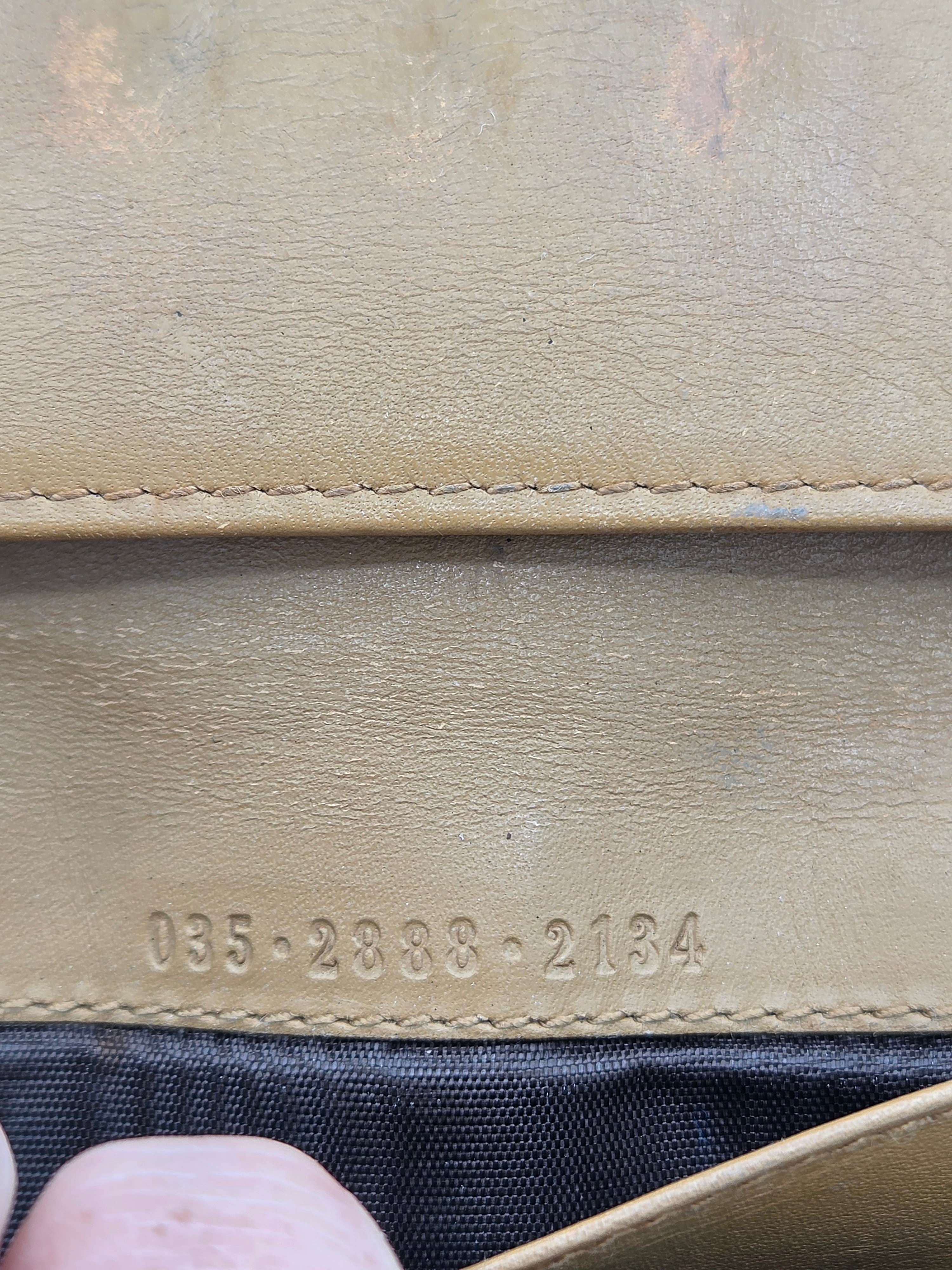 Close-up of a tan leather wallet or clutch showing the interior lining, stitched edge, embossed serial numbers, and a dark fabric interior.