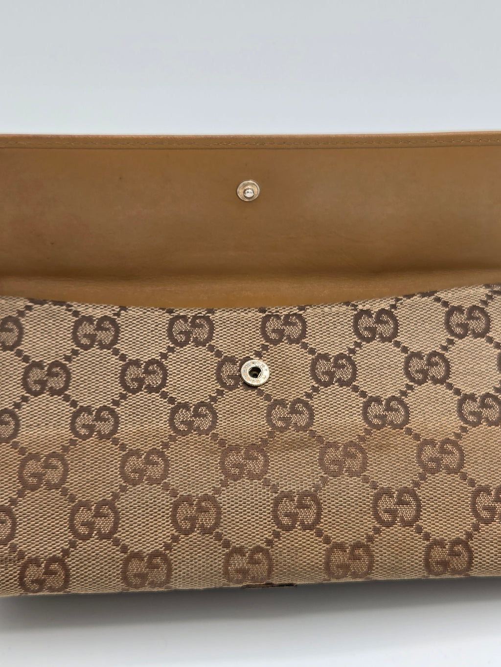 Close-up of a beige and brown monogram clutch or wallet with a tan leather strap and snap button closure, featuring the interlocking GG pattern.