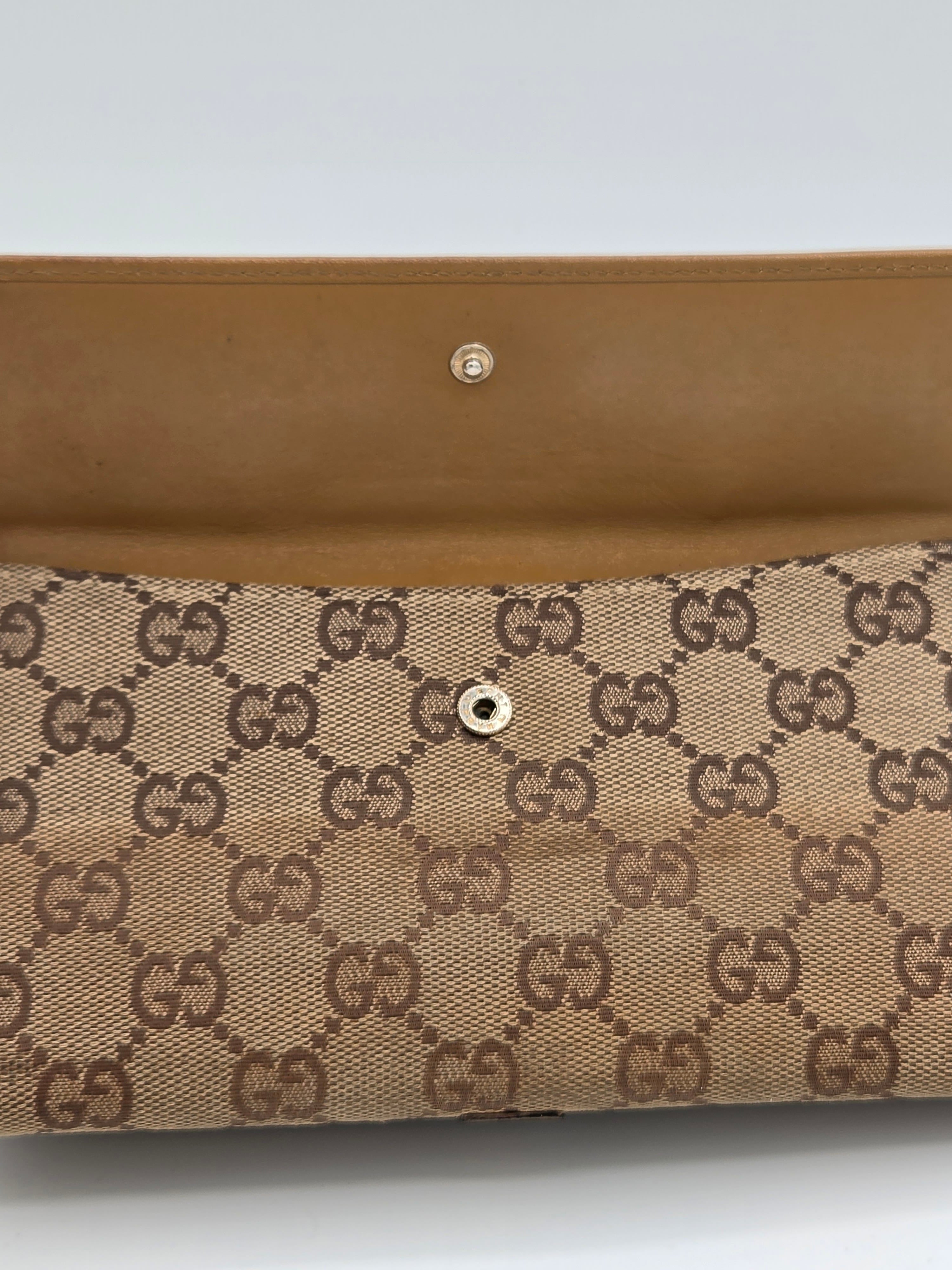 Close-up of a beige and brown monogram clutch or wallet with a tan leather strap and snap button closure, featuring the interlocking GG pattern.