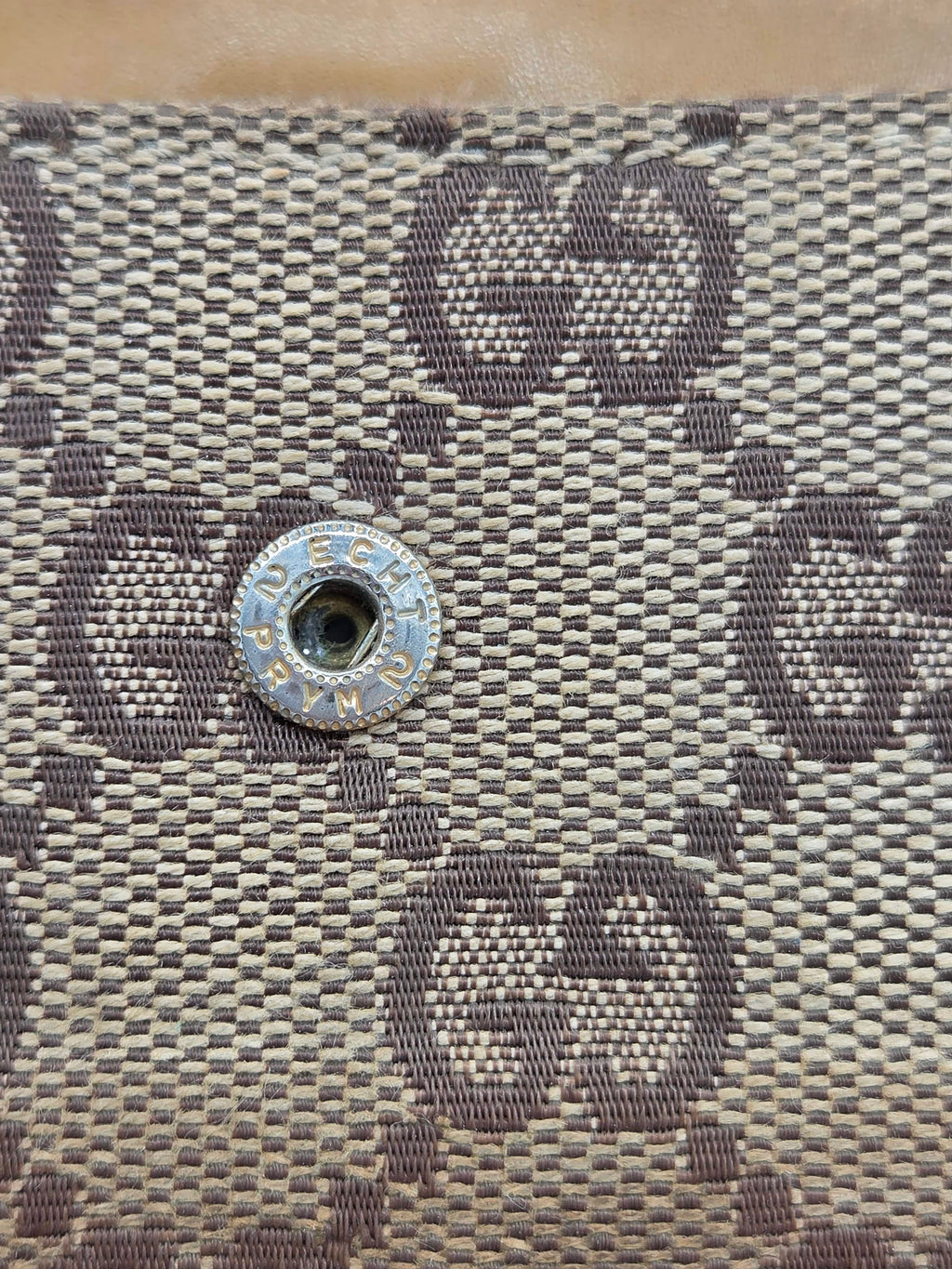 Close-up of a woven monogram fabric with a silver-tone metal snap button on a beige and gray background.