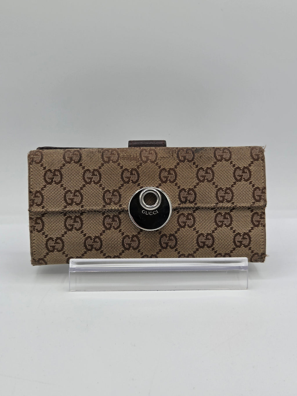 A brown and beige monogrammed Gucci pouch with a black circular snap button, shown next to a clear plastic sleeve on a light gray background.