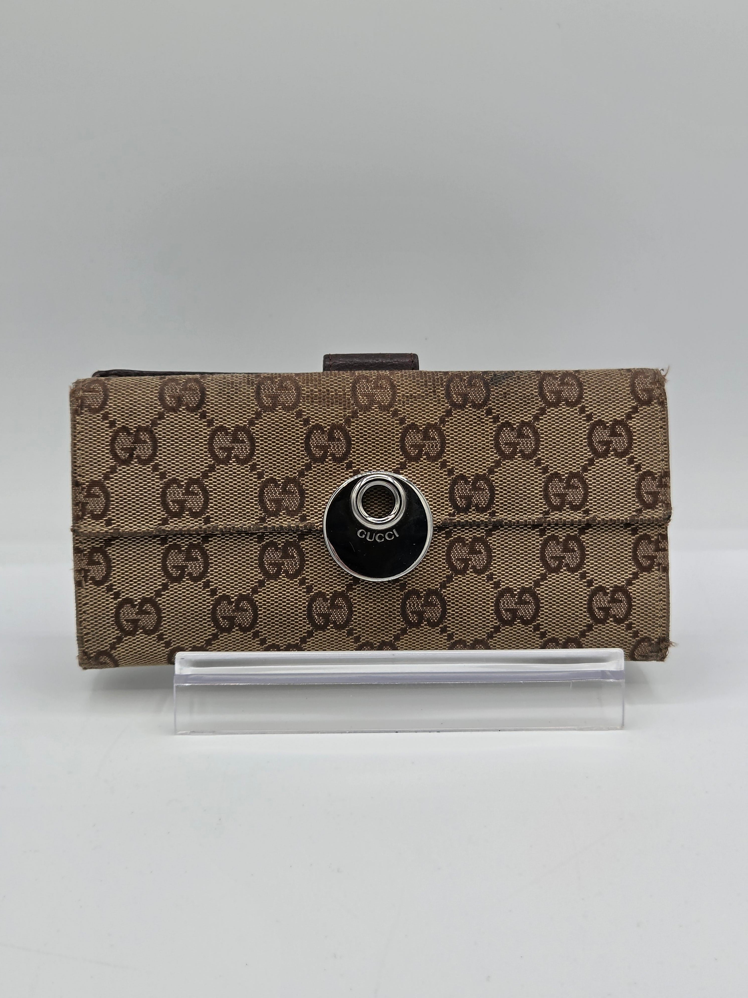 A brown and beige monogrammed Gucci pouch with a black circular snap button, shown next to a clear plastic sleeve on a light gray background.