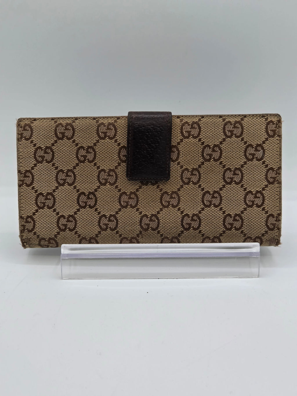 A Gucci monogram canvas notebook or planner cover with a dark brown leather strap closure, shown next to a clear plastic sleeve on a plain light background.
