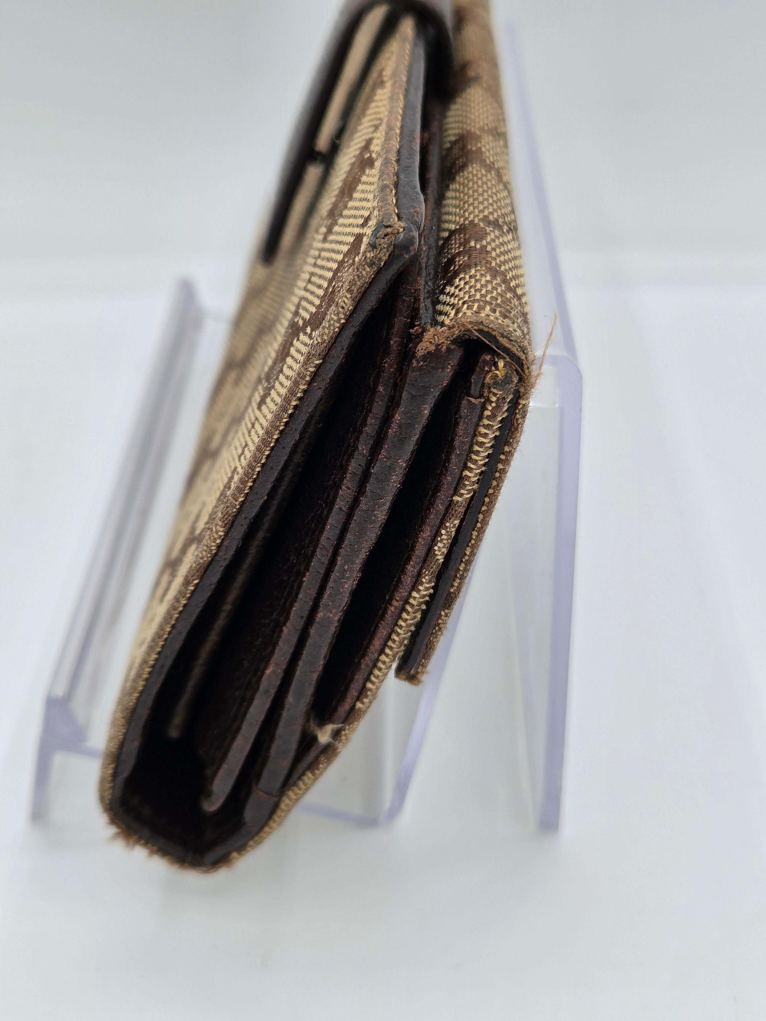 A close-up of an open bi-fold wallet with a tan and black woven patterned exterior and dark brown interior card slots, resting on a clear acrylic stand against a white background.