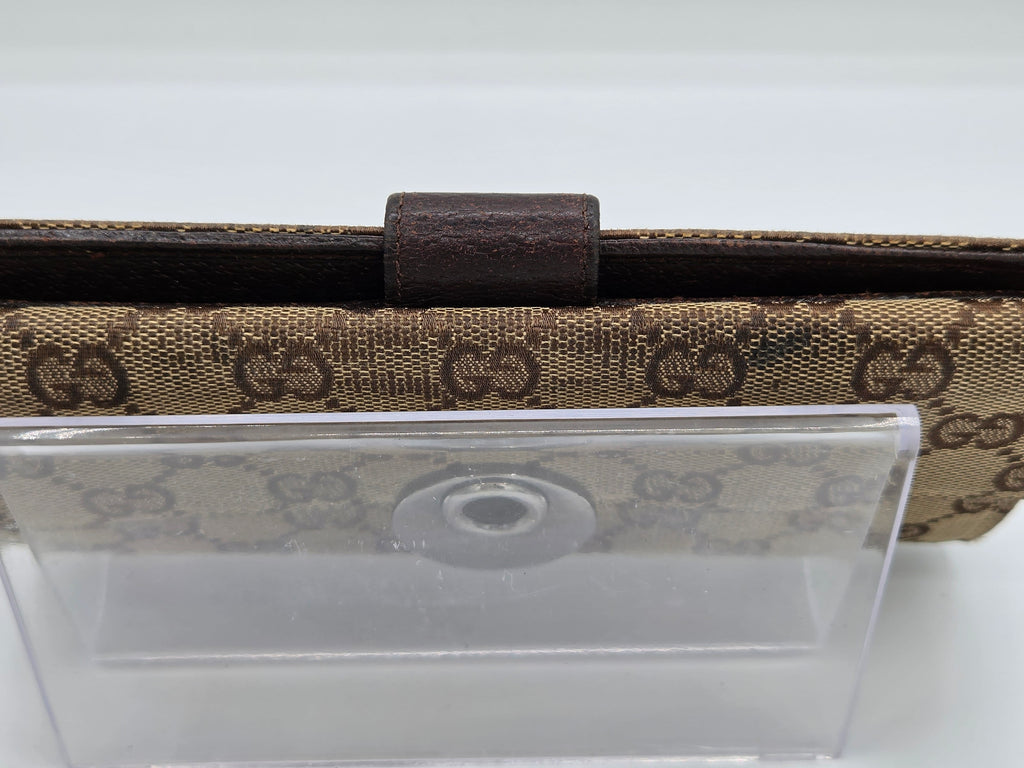A close-up of a Gucci wallet or clutch featuring the brand's signature GG monogram canvas in beige and brown, with a dark brown leather tab closure and a clear plastic protective cover resting on top.