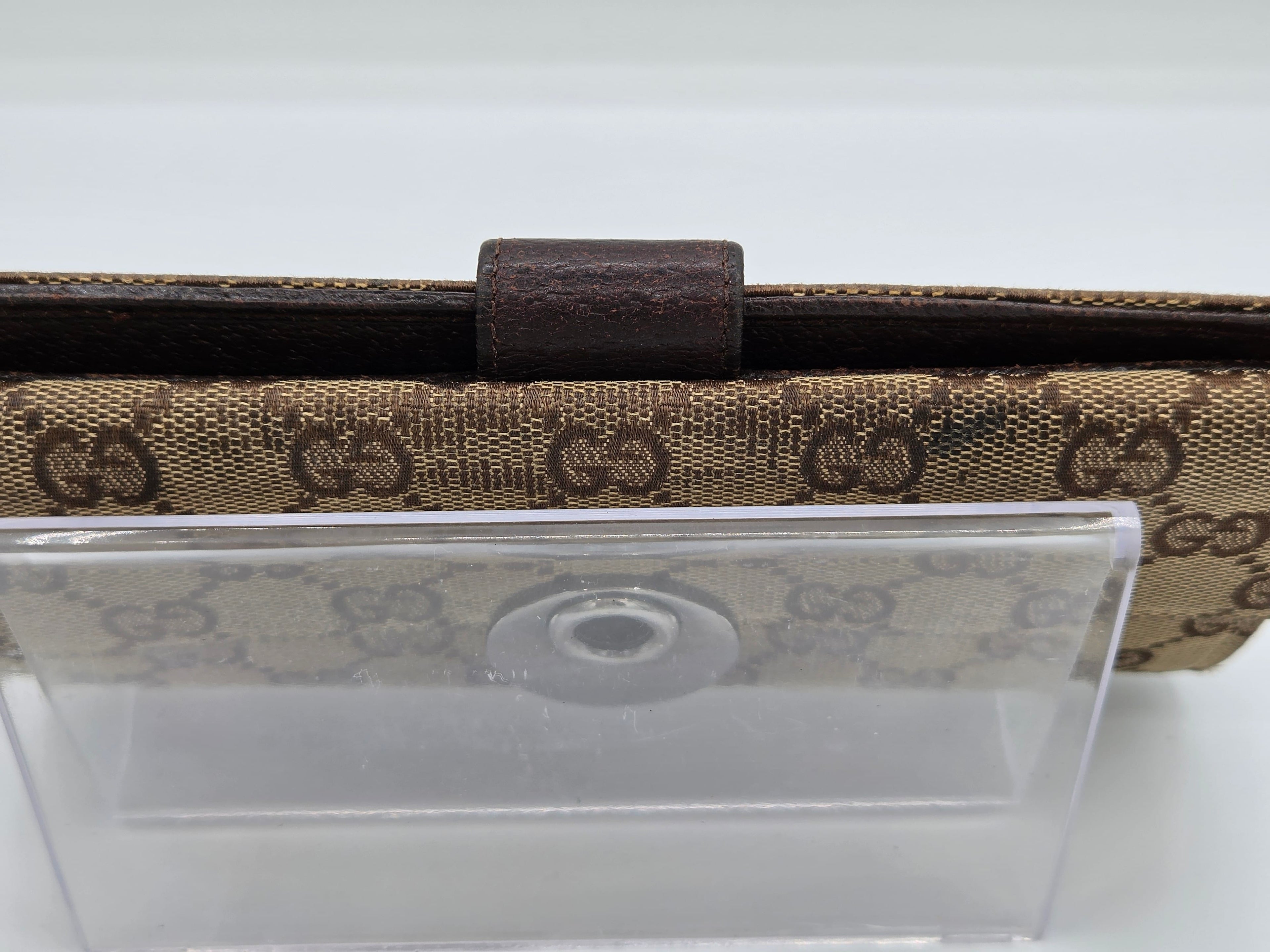 A close-up of a Gucci wallet or clutch featuring the brand's signature GG monogram canvas in beige and brown, with a dark brown leather tab closure and a clear plastic protective cover resting on top.