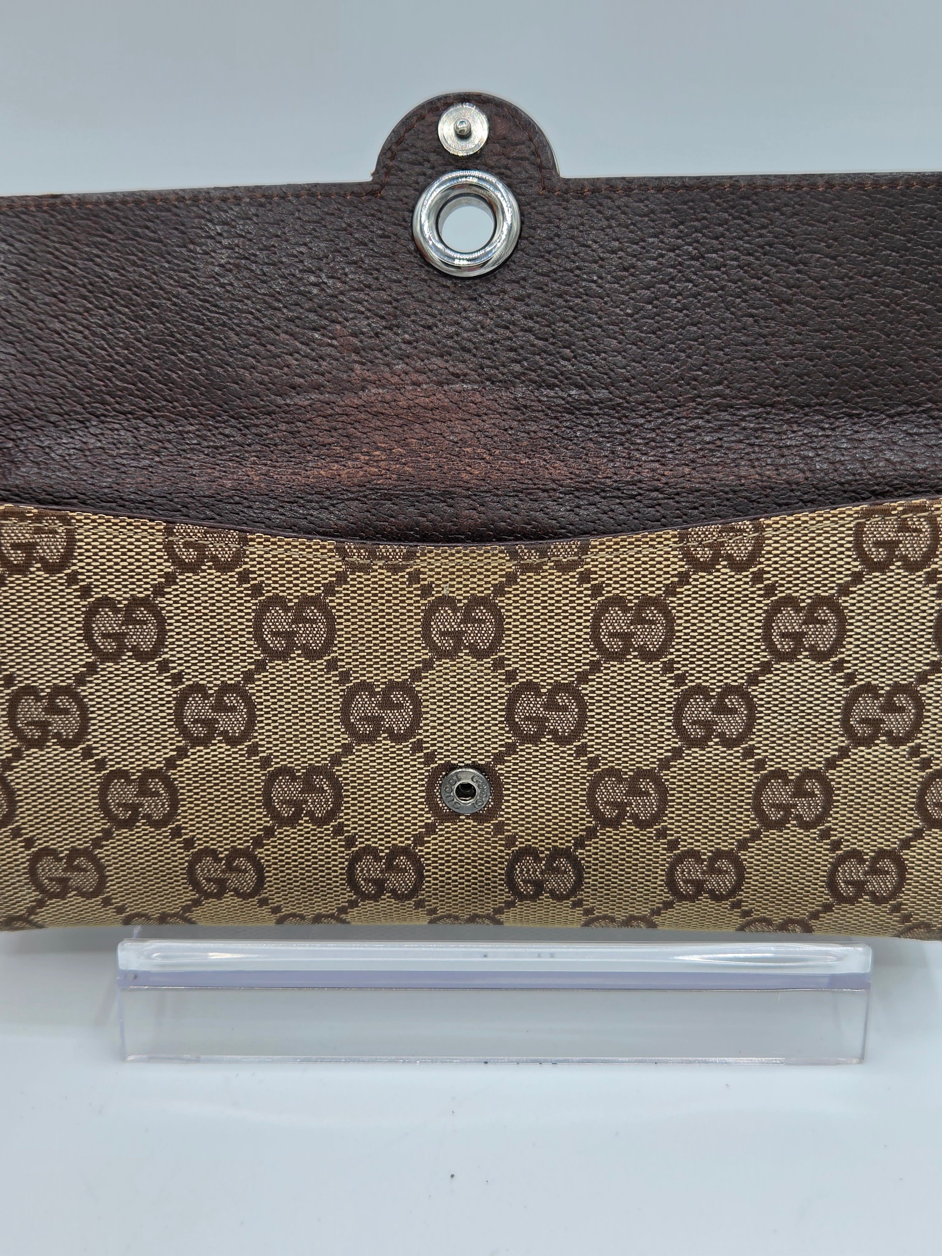 A close-up of a Gucci clutch or wallet featuring the classic brown and beige GG monogram canvas on the front flap and a dark brown leather top section with a silver-tone metal snap button.