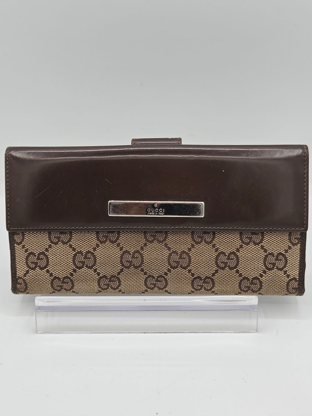 A Gucci clutch wallet featuring the brand's brown GG Supreme canvas pattern on the front cover and a solid dark brown leather side panel with a silver-tone metal nameplate.