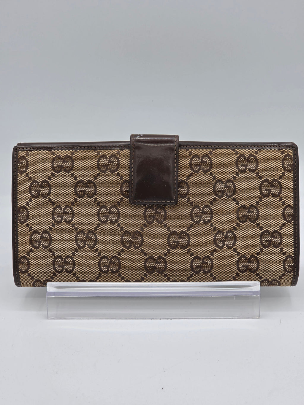 A brown and beige monogram wallet with a dark brown leather strap closure, shown next to a clear plastic sleeve on a light gray background.