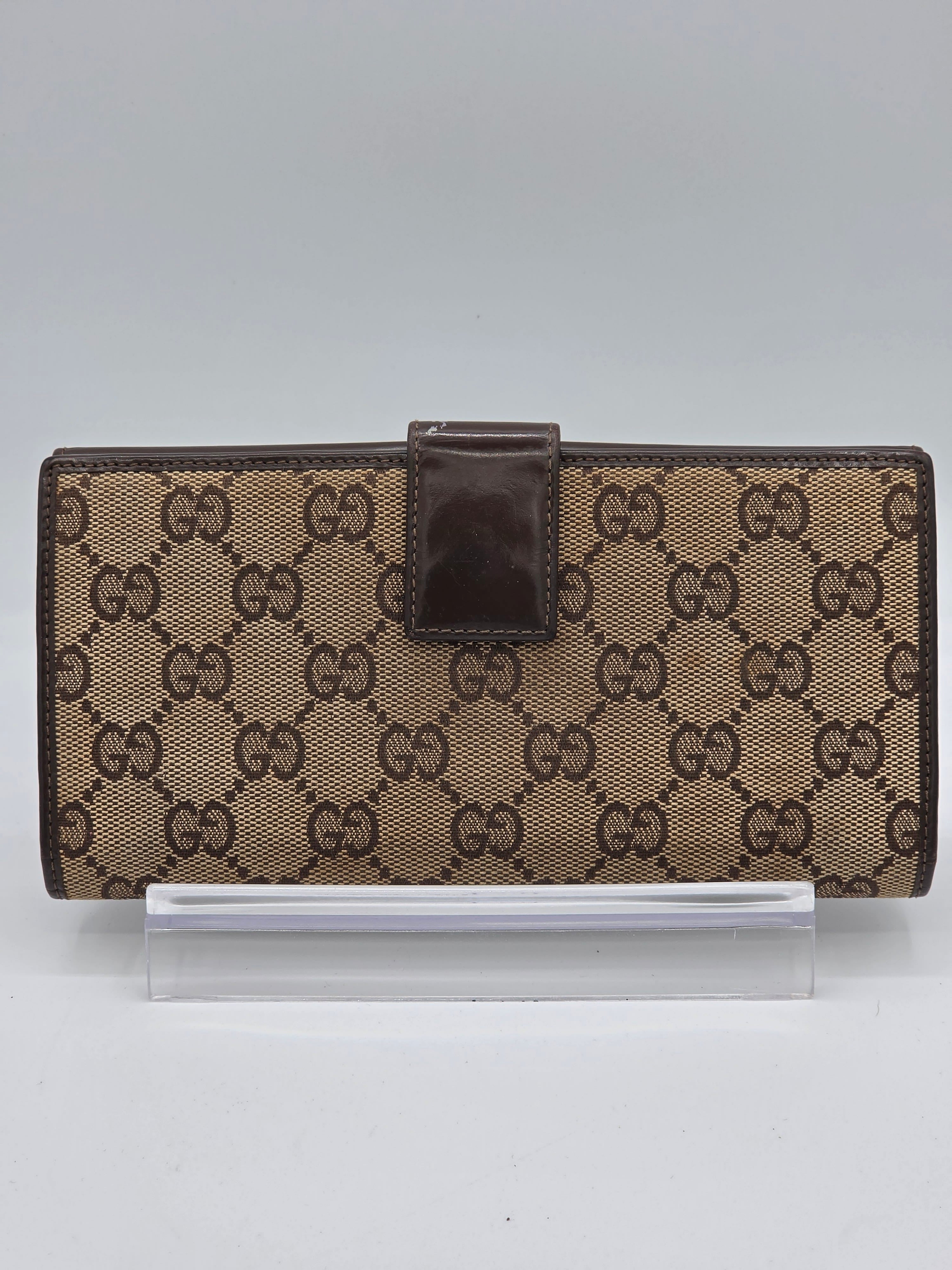 A brown and beige monogram wallet with a dark brown leather strap closure, shown next to a clear plastic sleeve on a light gray background.