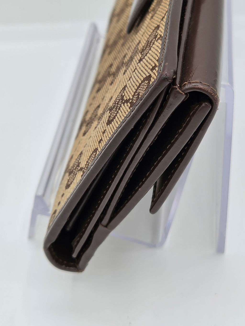 A close-up of a brown leather wallet with a beige and brown monogram patterned exterior, resting on a clear acrylic display stand against a white background.