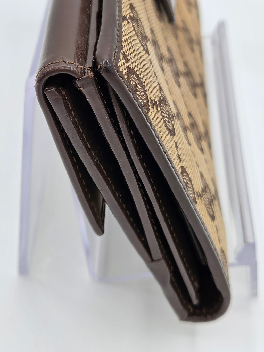 A close-up of a brown and beige patterned leather wallet resting on a clear acrylic display stand against a white background.