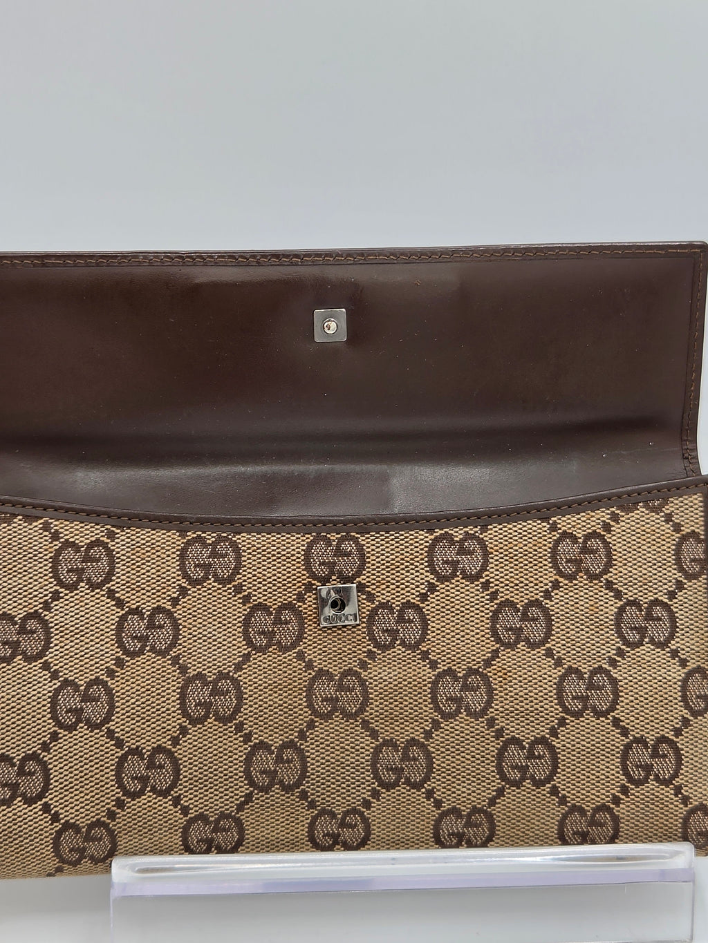 A close-up of a brown leather and beige GG Supreme canvas clutch or wallet, showing the interior flap with a silver-tone snap button and the exterior patterned side.