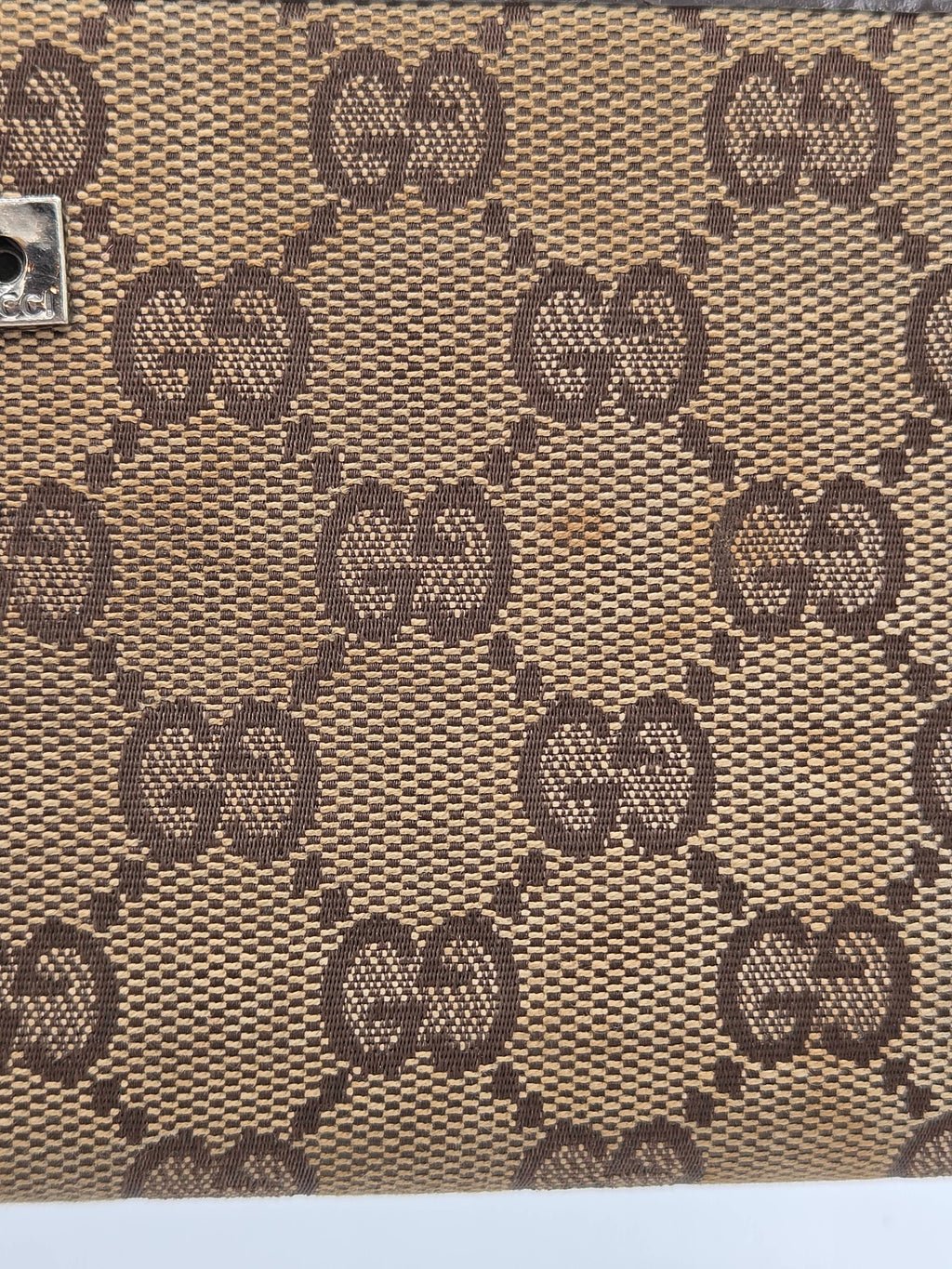 Close-up of a beige and brown monogram patterned material with repeating interlocking GG motifs, likely coated canvas or fabric.