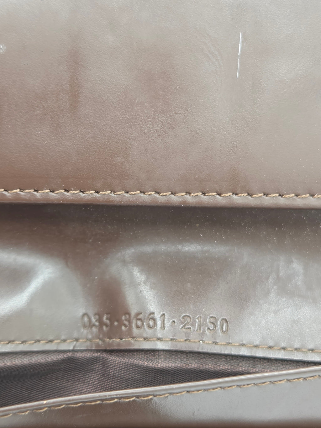 Close-up detail of a taupe-brown leather or faux-leather product, showing a stitched seam, a glossy panel with embossed serial numbers, and a dark mesh-like section on the right.