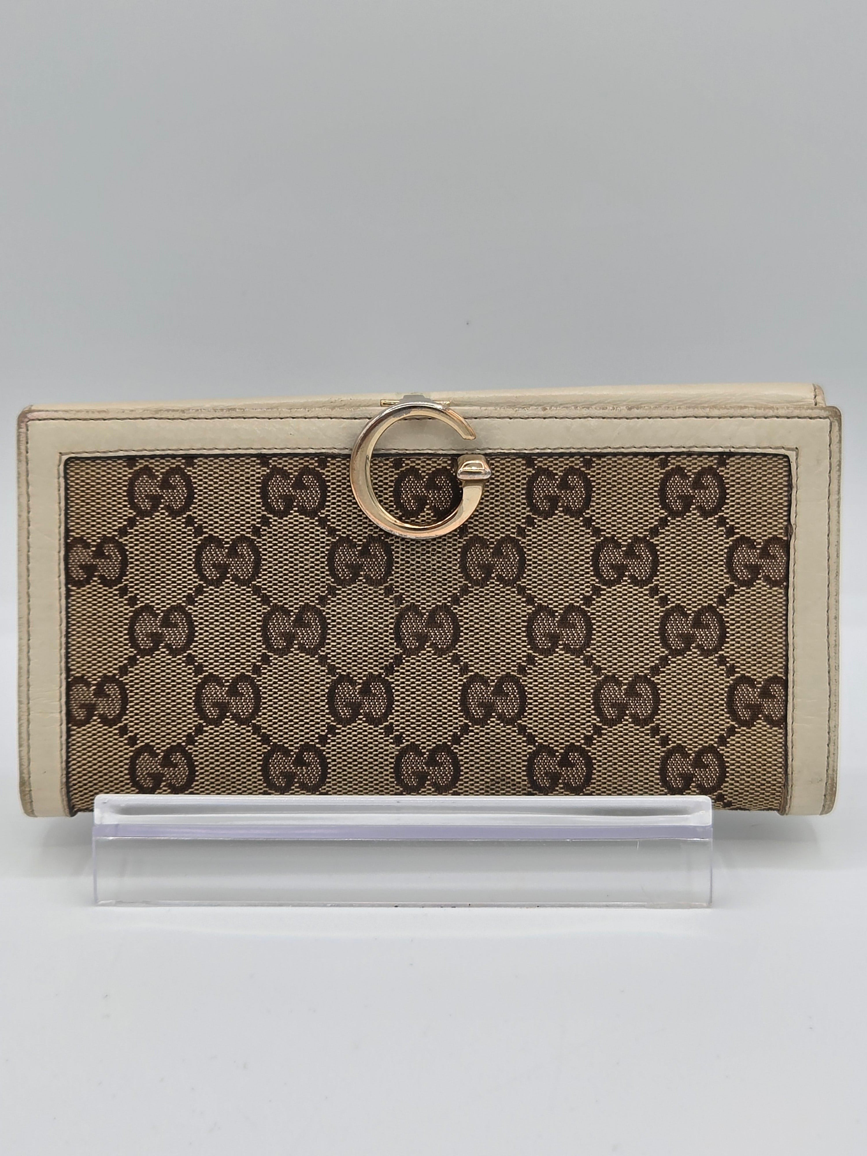 A vintage-style Gucci monogram wallet in beige and brown canvas with a gold-tone ring pull, shown next to a clear plastic sleeve on a light gray background.