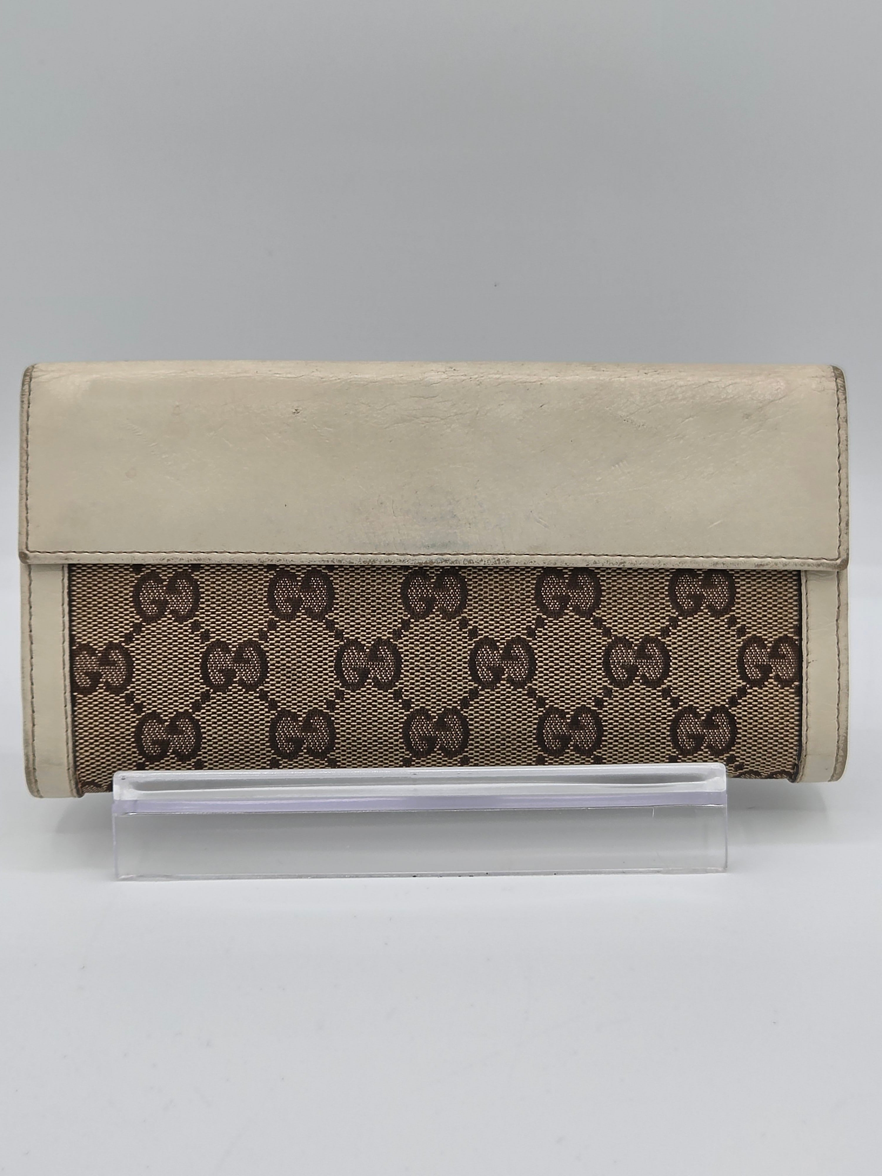 A Gucci monogram canvas and leather wallet in beige and brown, displayed on a clear acrylic stand against a light gray background.