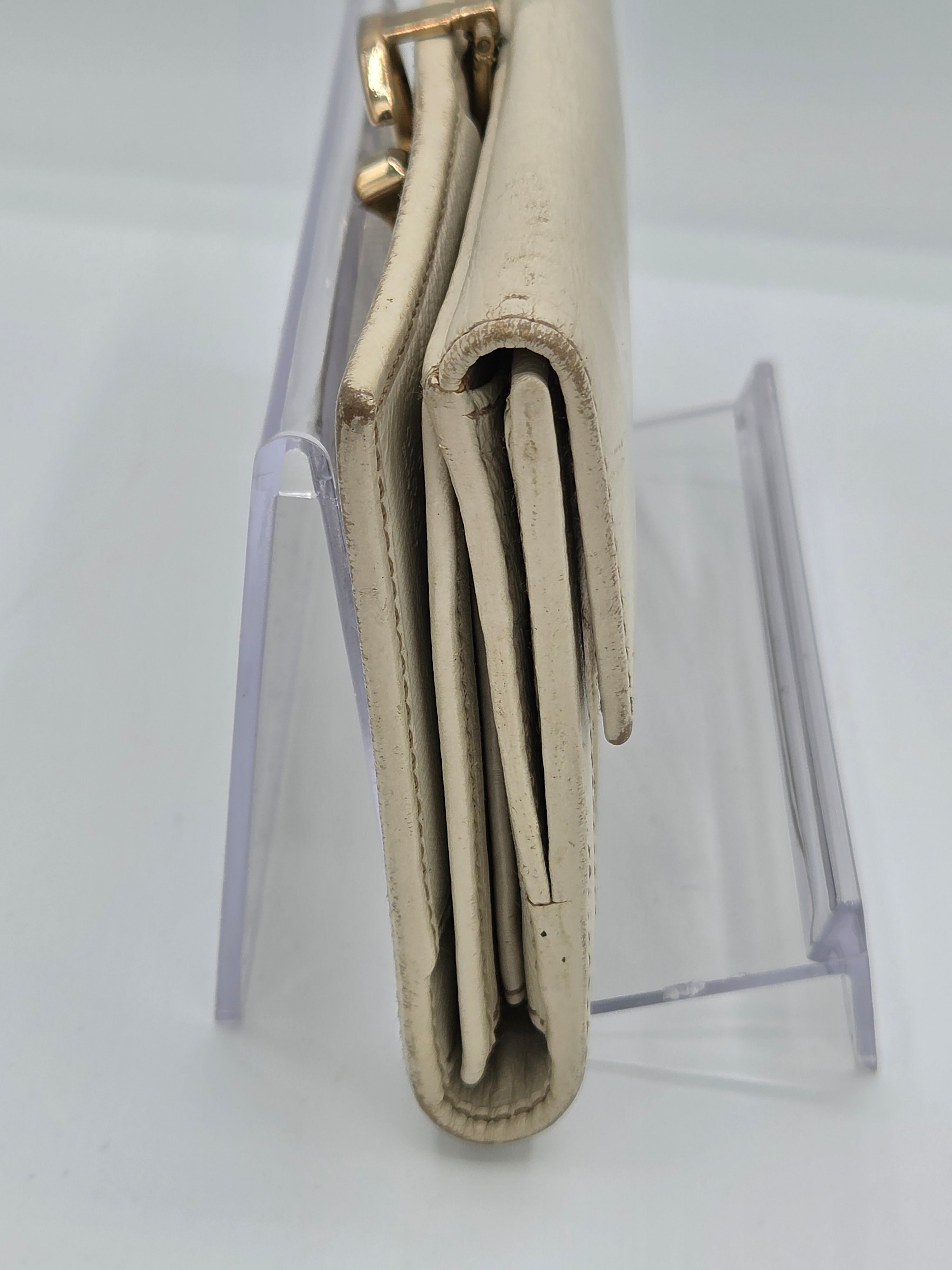 A folded beige leather wallet or clutch with gold-tone hardware, displayed horizontally on a clear acrylic stand against a white background.