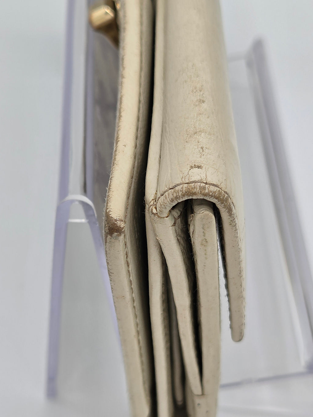 A close-up of a light beige leather wallet or clutch showing the rolled edge and interior fold, resting on a clear acrylic stand against a white background.