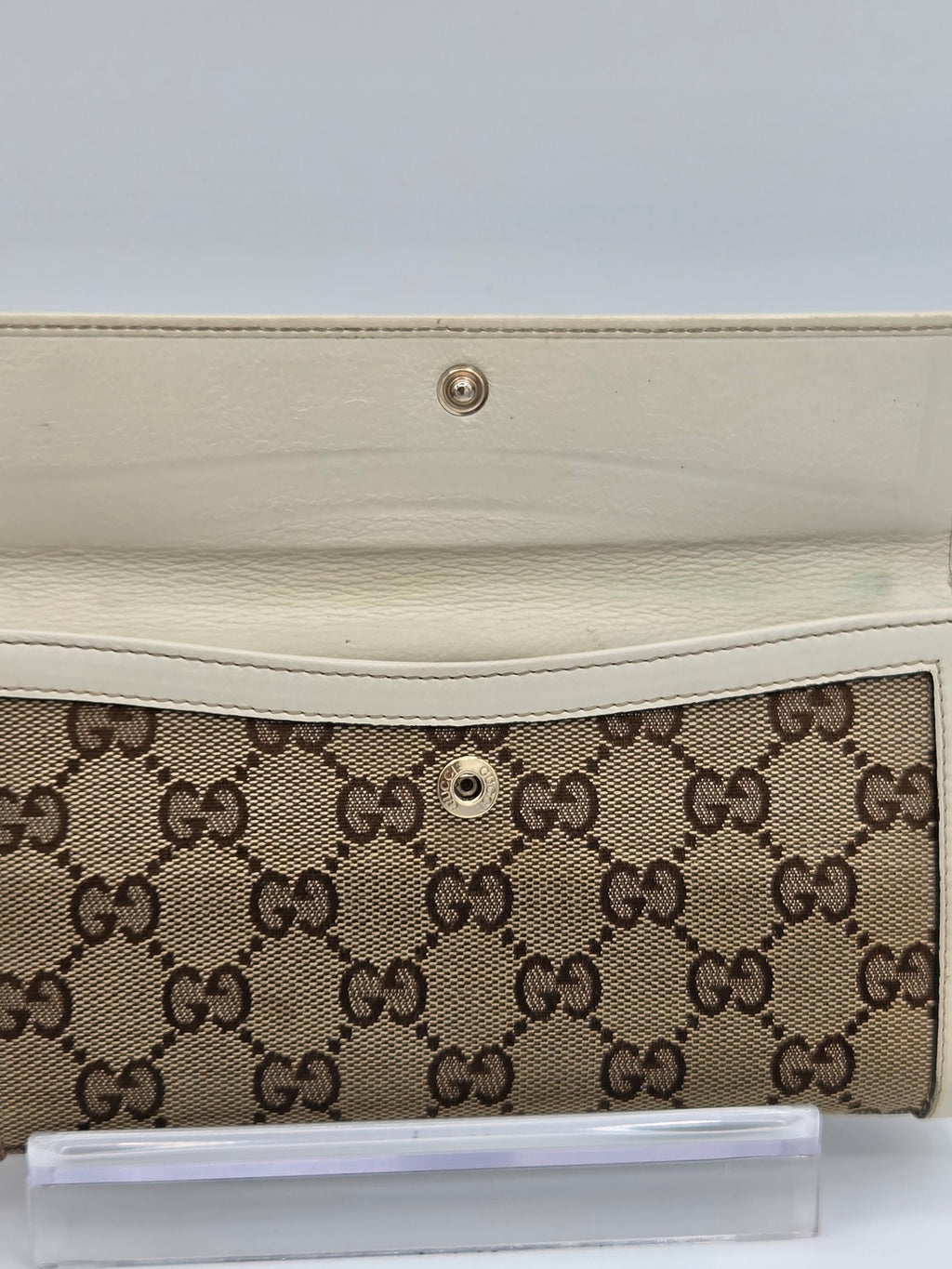 A close-up of a Gucci wallet or clutch featuring the brand's signature GG monogram canvas in beige and brown, with a cream-colored leather flap and a gold-tone snap button.