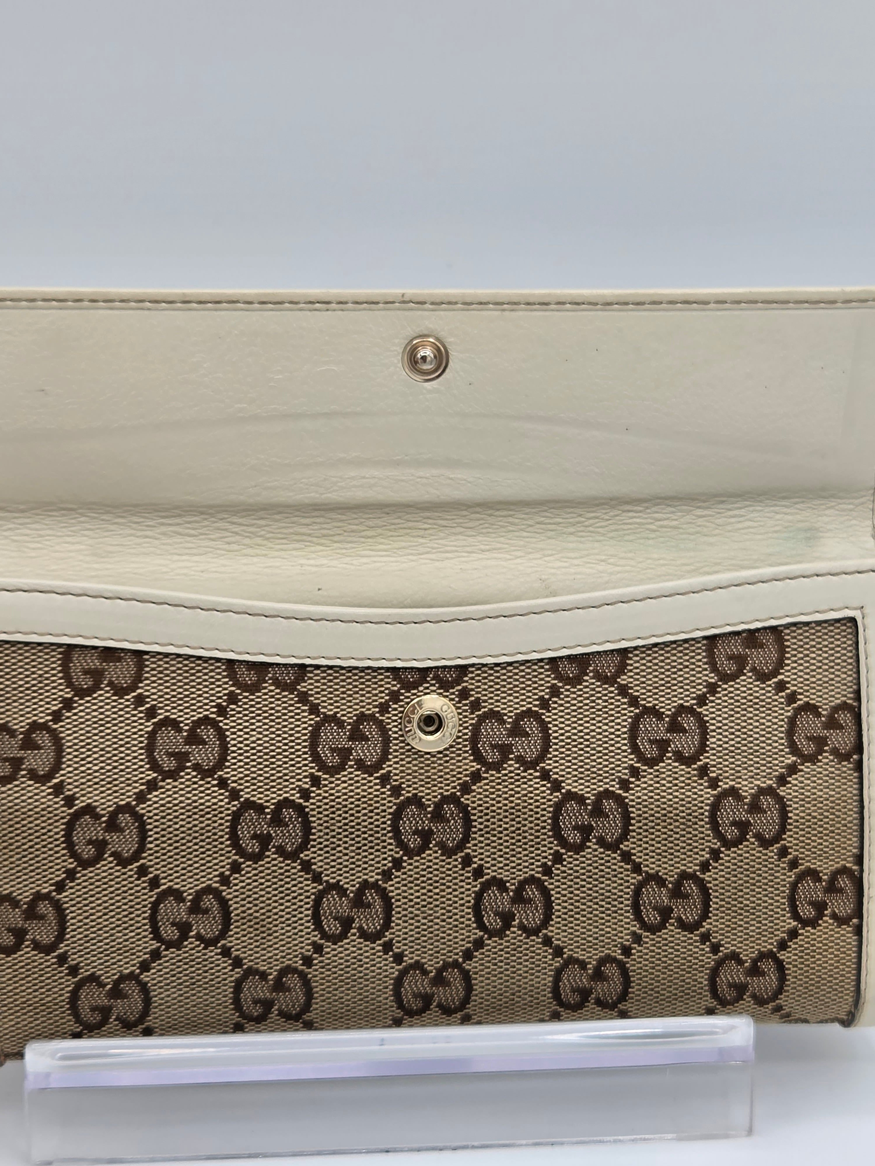 A close-up of a Gucci wallet or clutch featuring the brand's signature GG monogram canvas in beige and brown, with a cream-colored leather flap and a gold-tone snap button.