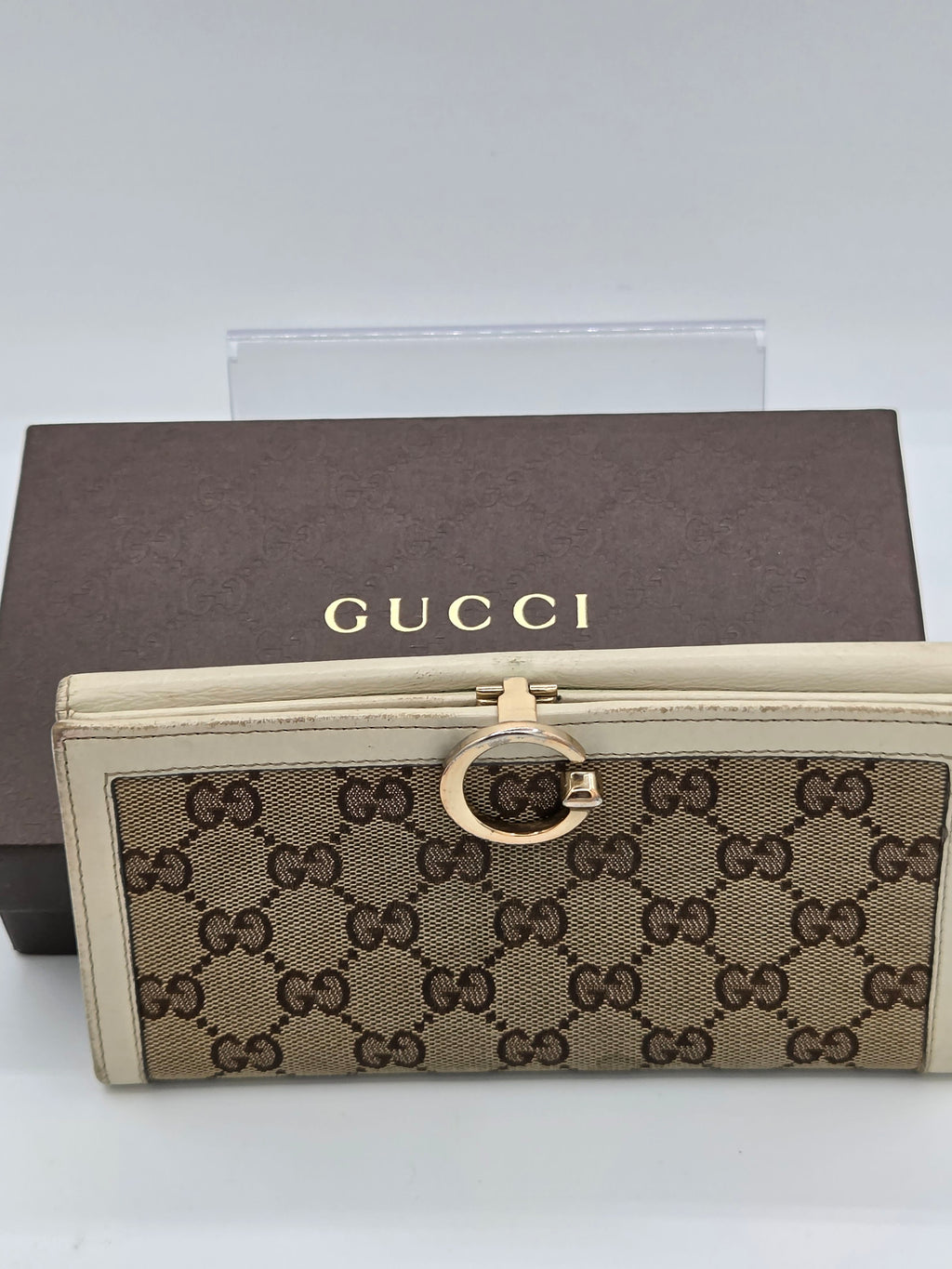A Gucci monogram clutch wallet with a gold-tone G-ring clasp, displayed in front of its brown Gucci box on a light gray background.