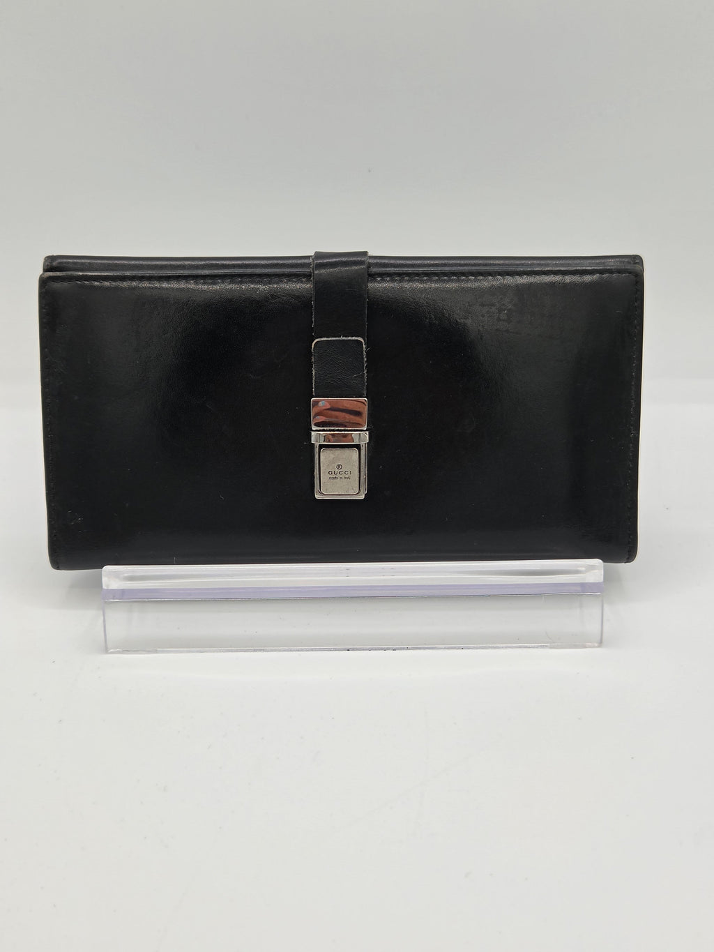 A black leather clutch wallet with a silver-toned metal clasp, displayed on a clear acrylic stand against a light gray background.