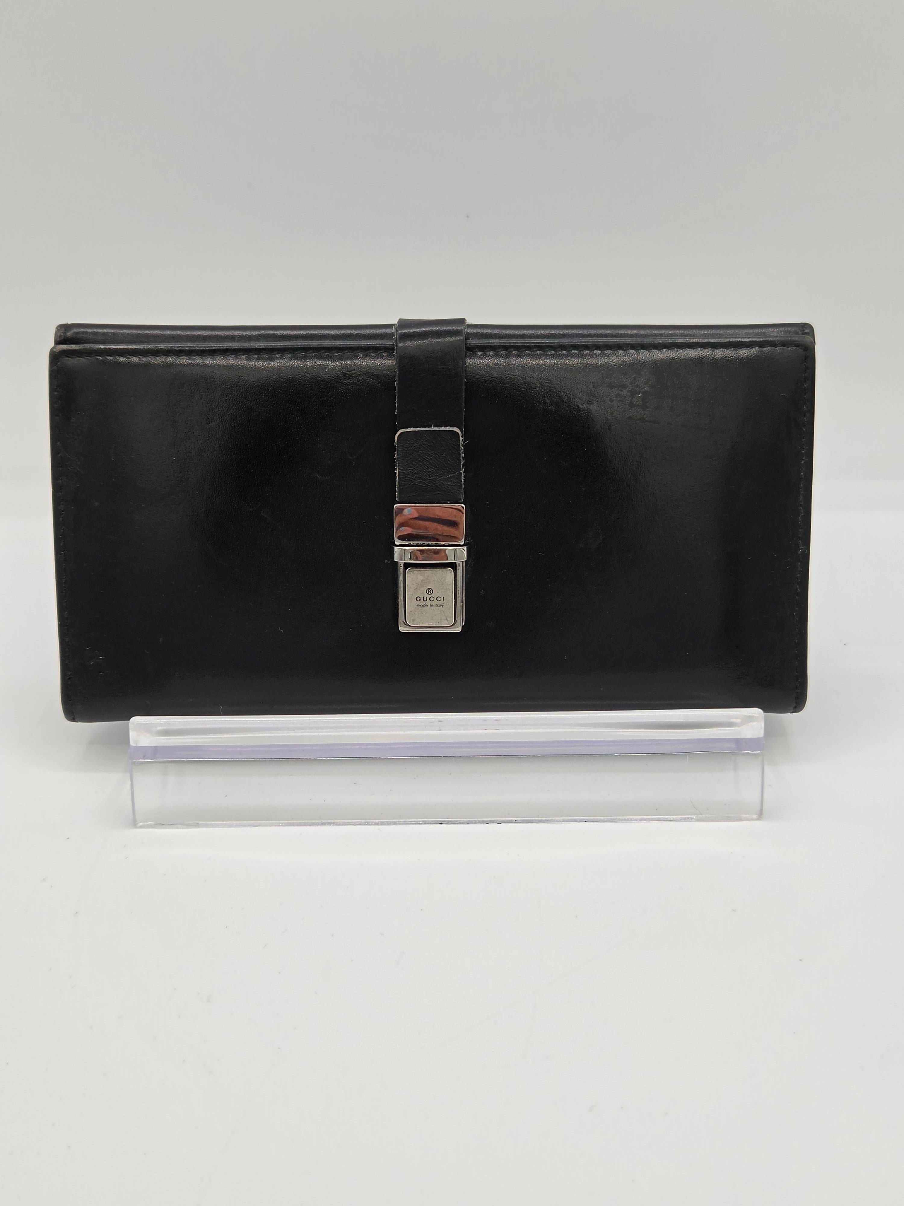 A black leather clutch wallet with a silver-toned metal clasp, displayed on a clear acrylic stand against a light gray background.