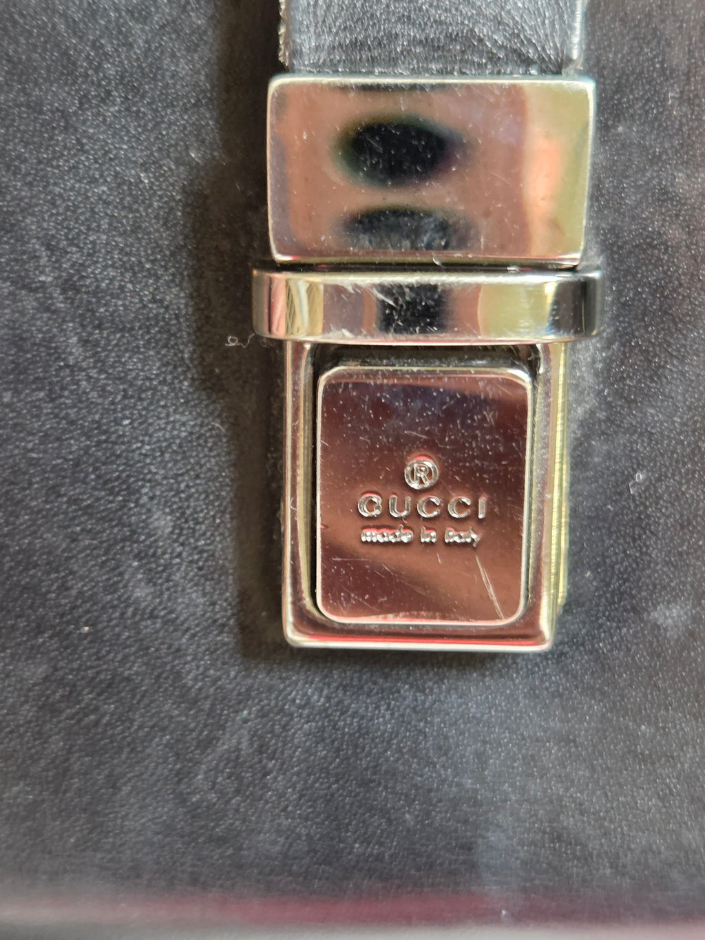 Close-up of a Gucci belt buckle with a polished gold-tone metal clasp engraved with the Gucci logo and 'made in Italy' on a dark textured background.