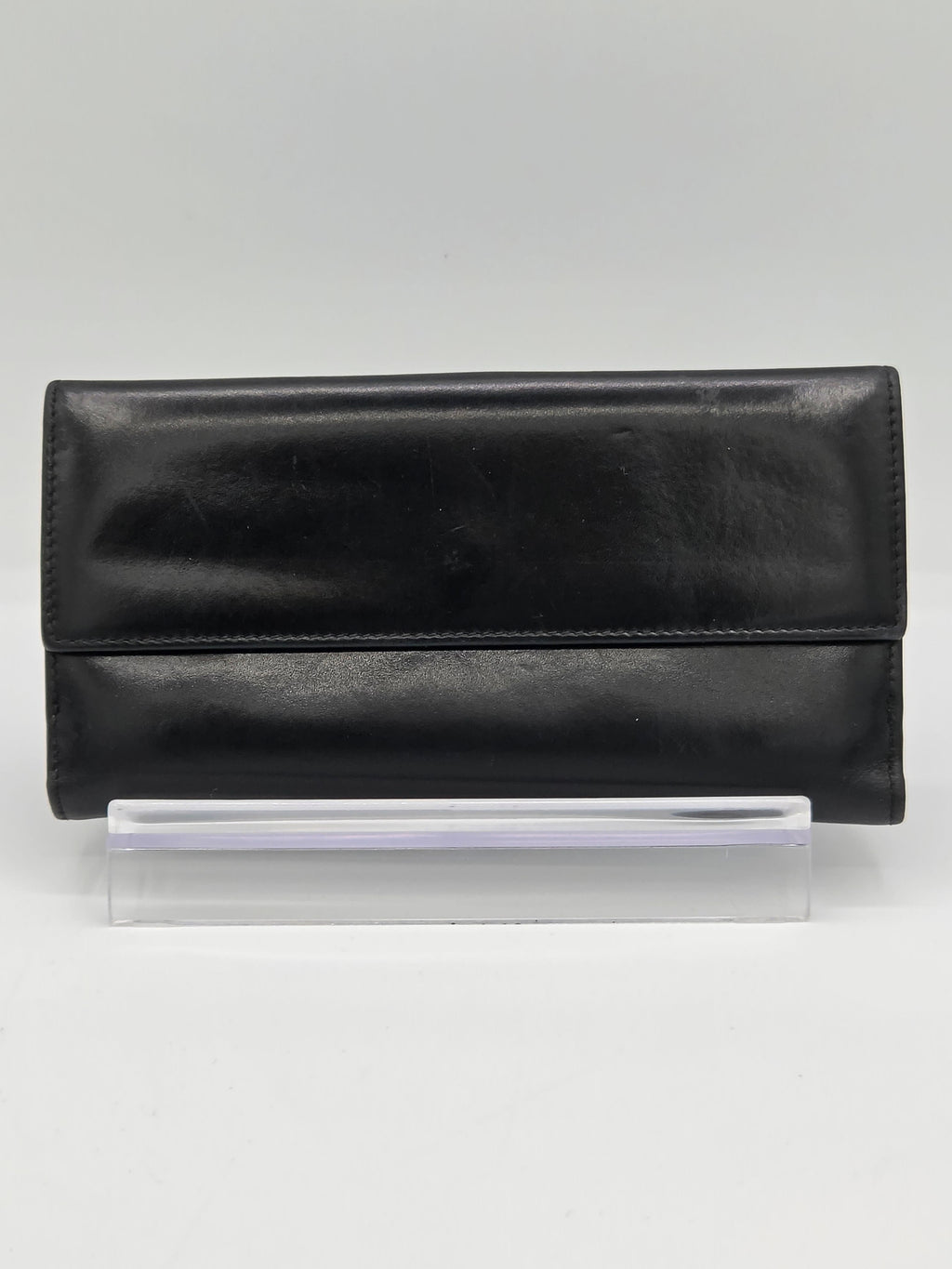 A black leather fold-over wallet or clutch displayed upright on a clear acrylic stand against a light gray background.