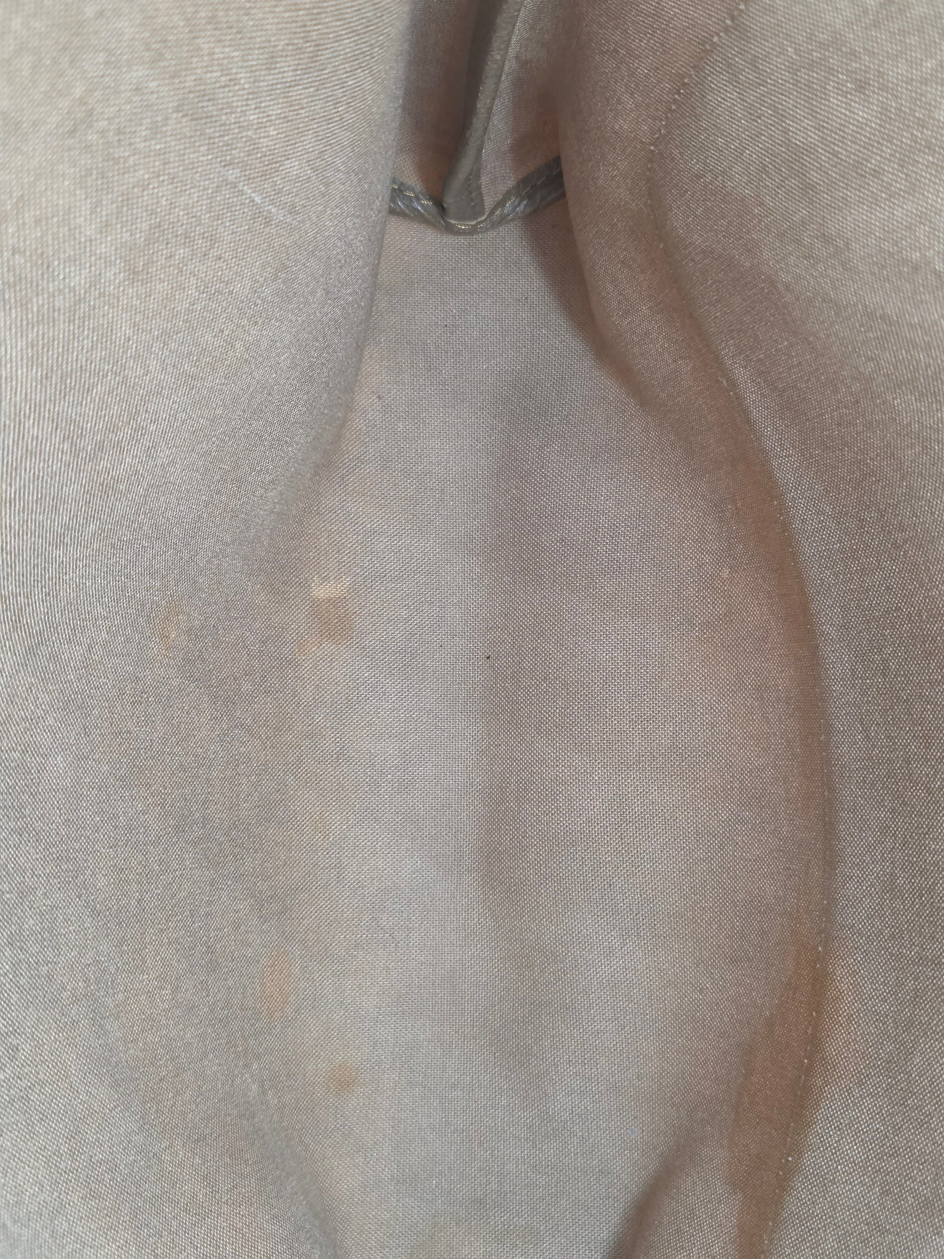 A close-up of a light beige or off-white textured fabric surface with soft folds and a small metallic loop visible on the left side.