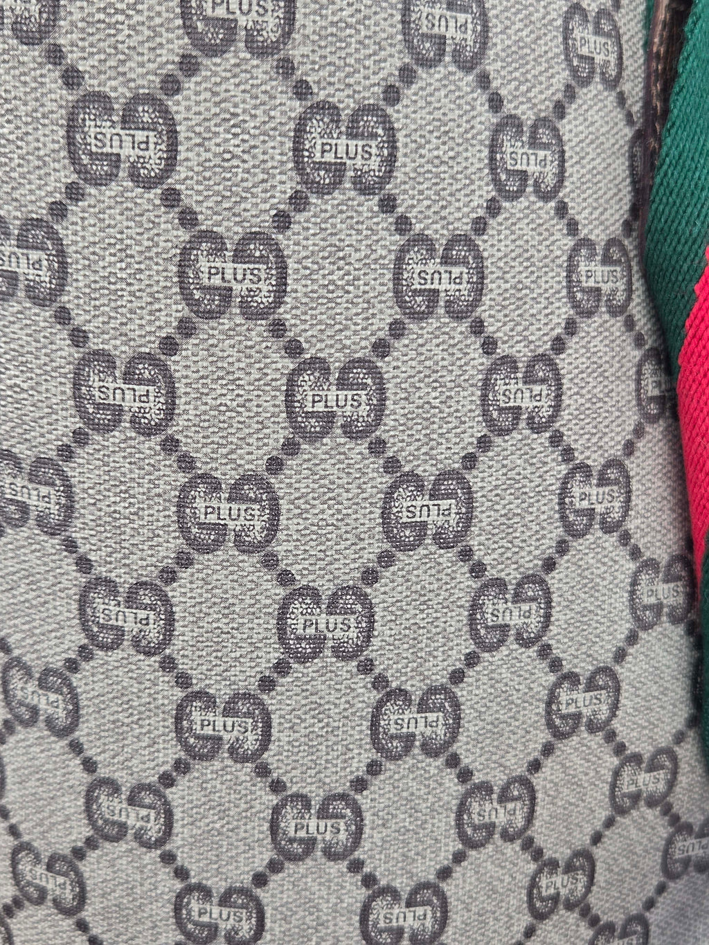 Close-up of a woven fabric with a repeating interlocking GG monogram pattern in gray and black, with partial green and pink fabric visible at the right edge.