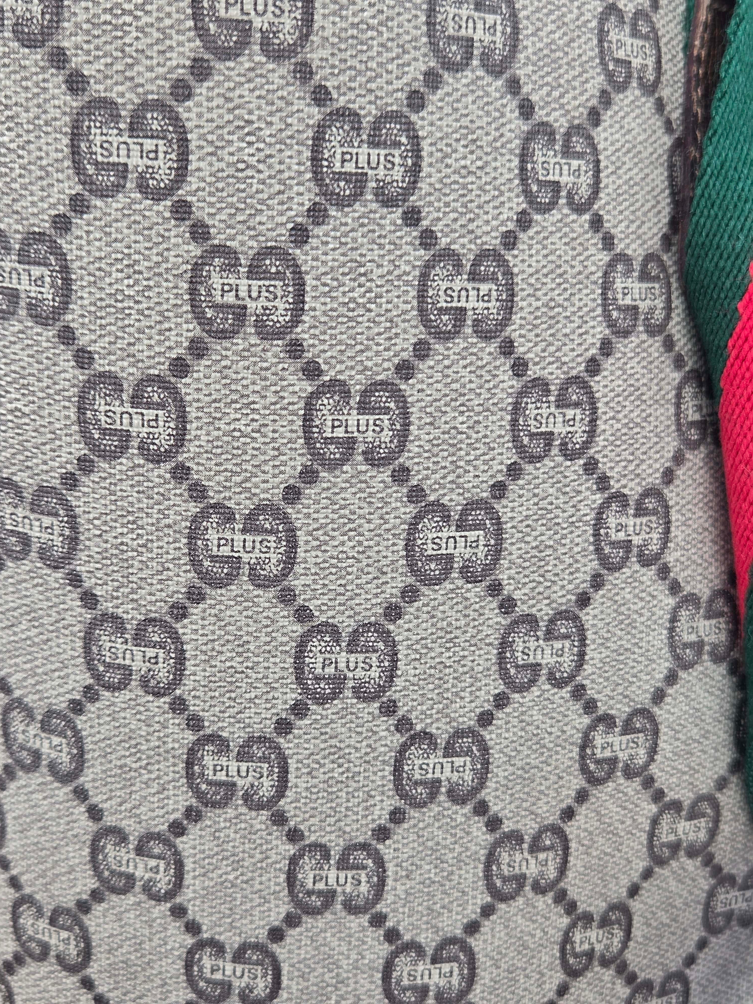 Close-up of a woven fabric with a repeating interlocking GG monogram pattern in gray and black, with partial green and pink fabric visible at the right edge.