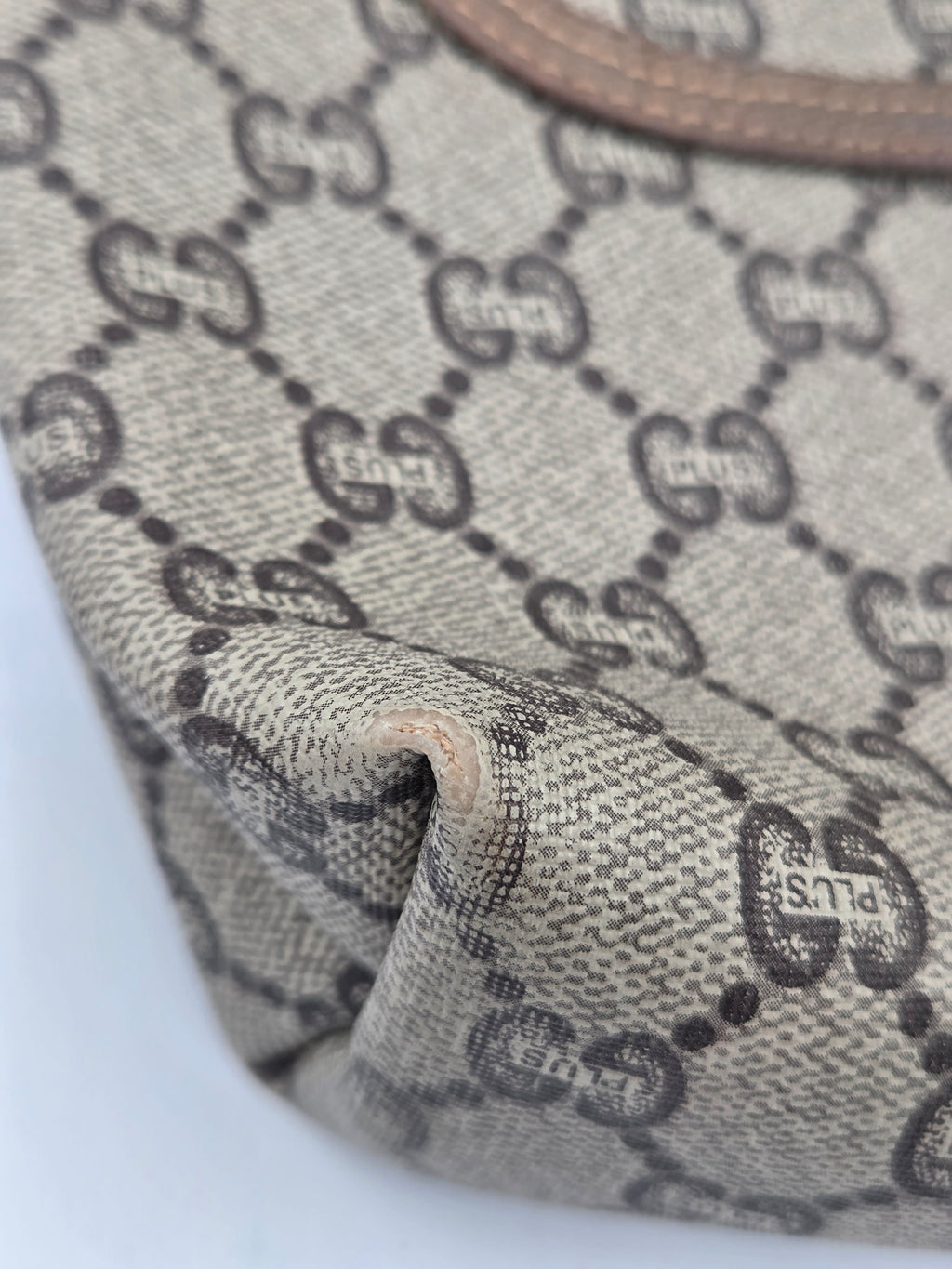 Close-up of a beige and gray monogram canvas material with a repeating interlocking G pattern and a small visible 'G' mark, showing fabric texture and a fold.