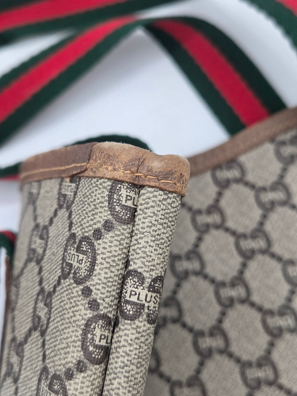 Close-up of a Gucci monogram canvas item with brown leather trim and a red and green striped ribbon detail, showing the classic GG pattern and a visible 'Gucci Plus' print on the fabric.