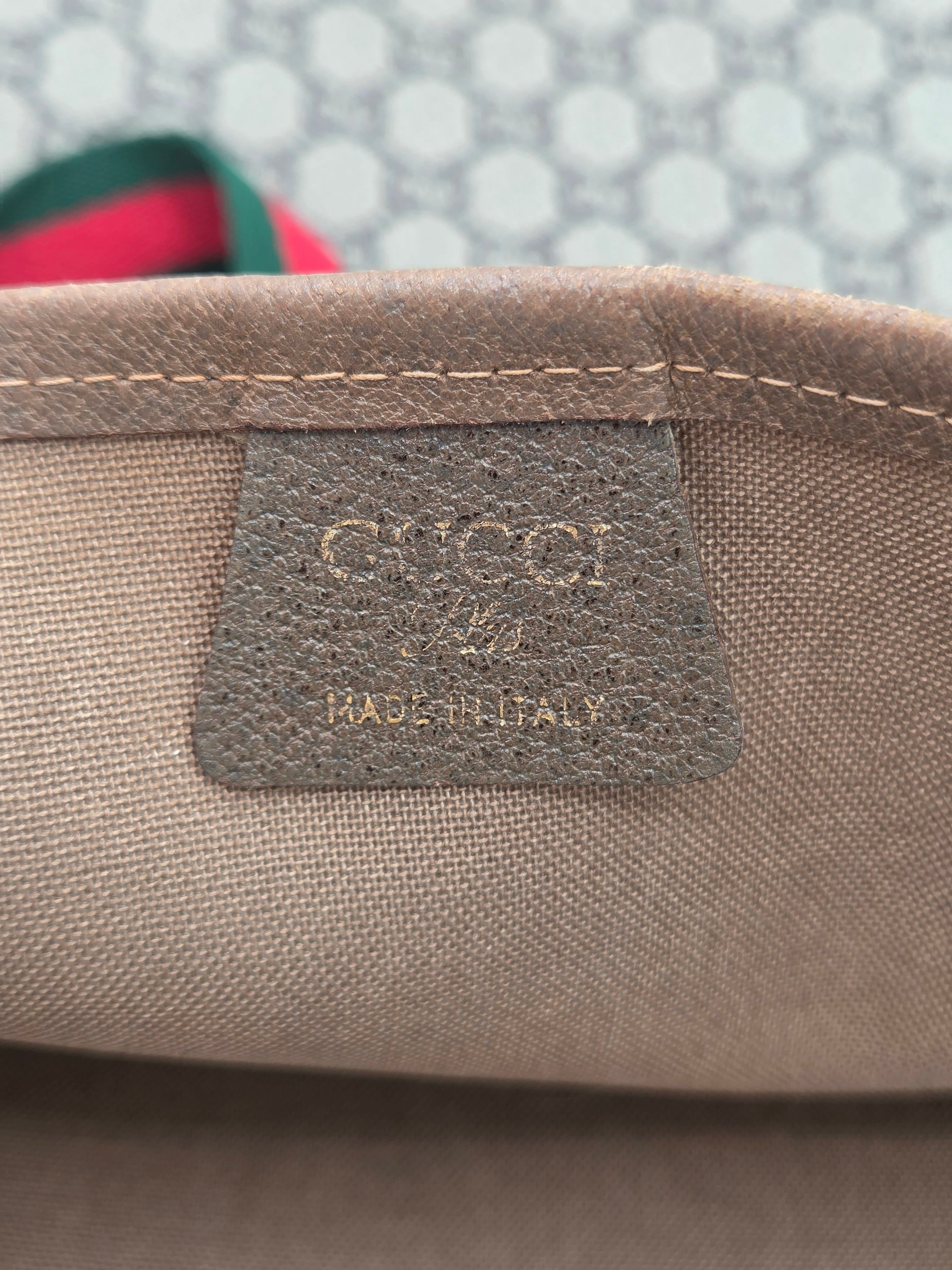 Close-up of a beige canvas bag corner showing a dark brown Gucci leather patch with embossed branding and 'MADE IN ITALY' text.