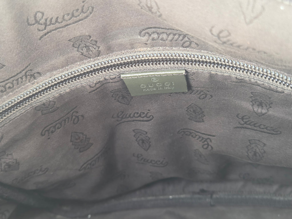 Close-up of the interior lining of a gray Gucci bag, showing a silver zipper, a metal Gucci brand tag, and an all-over embossed Gucci logo pattern.