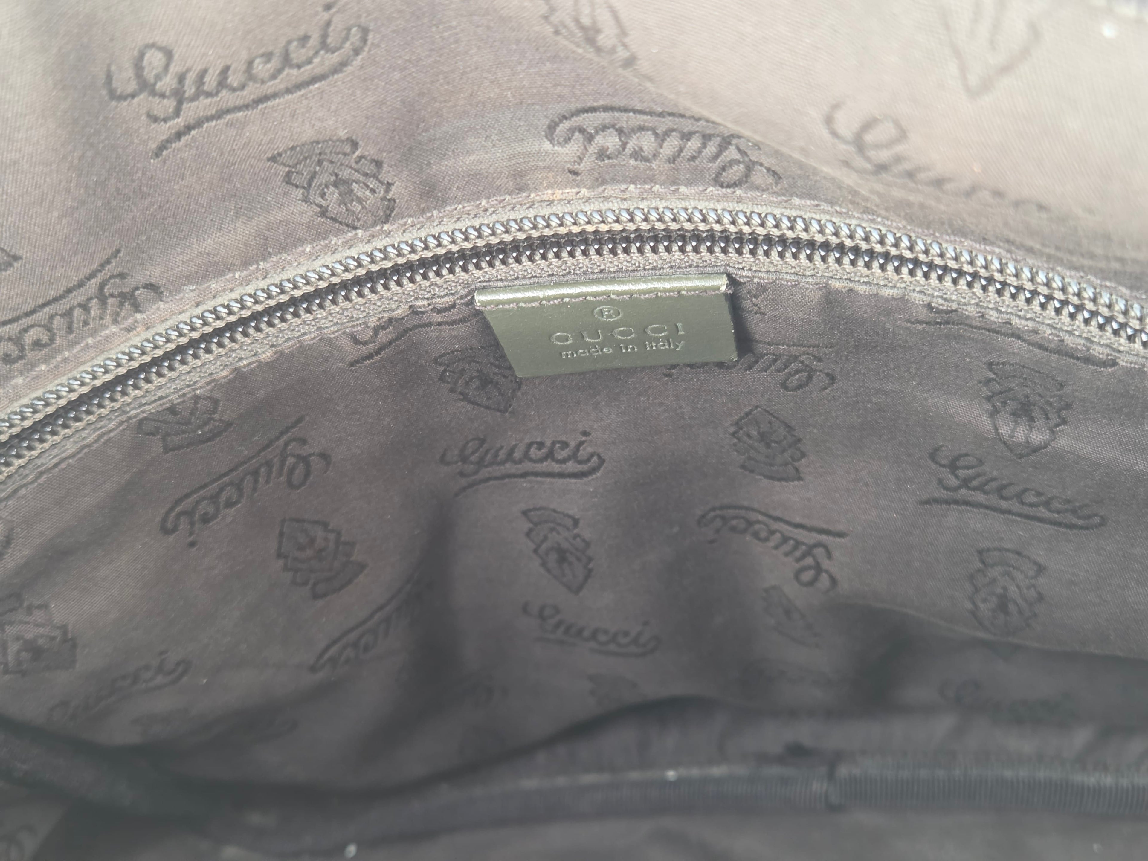 Close-up of the interior lining of a gray Gucci bag, showing a silver zipper, a metal Gucci brand tag, and an all-over embossed Gucci logo pattern.