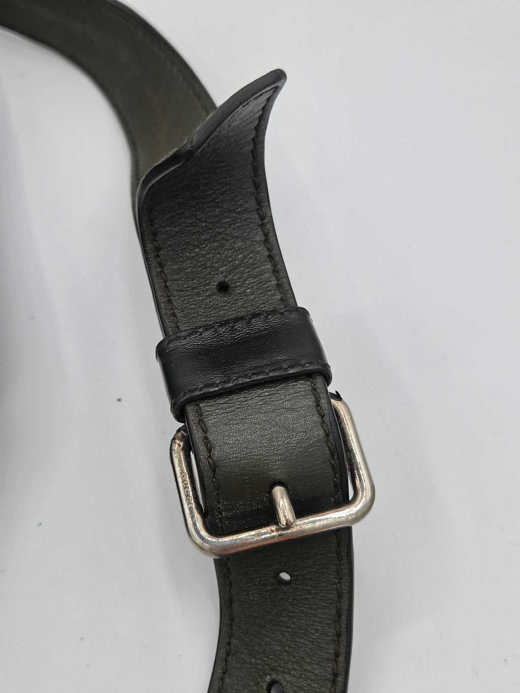 A close-up of a black leather belt with a silver-tone metal buckle, laid out on a plain light gray background.