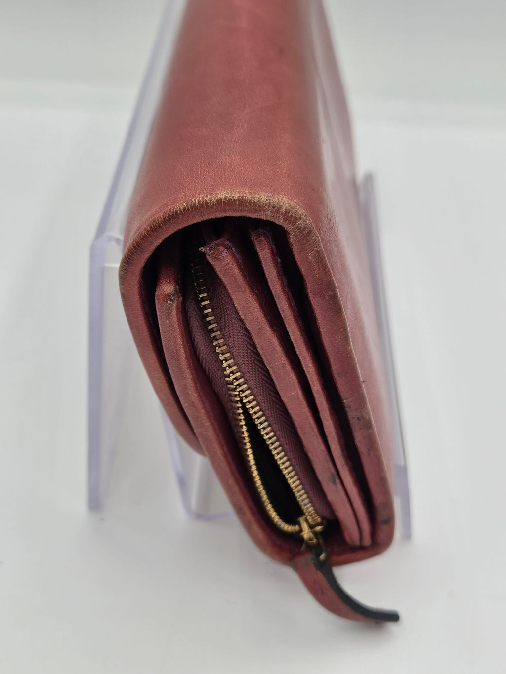 A close-up of a brown leather wallet or card holder with a gold zipper, resting on a clear acrylic stand against a white background.