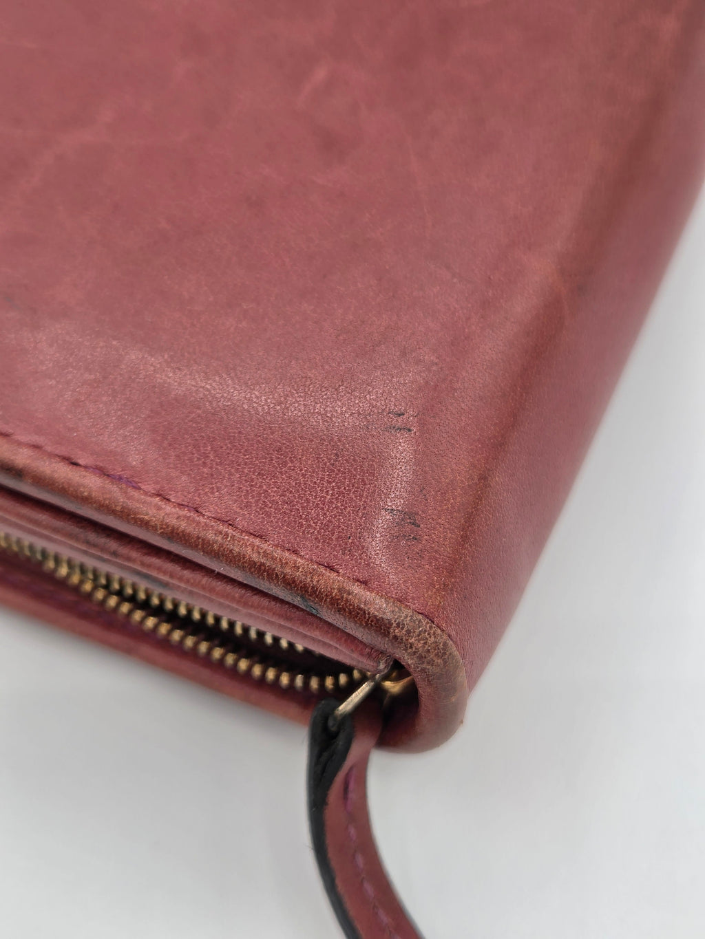 A close-up of a dusty rose leather clutch or wallet showing the textured leather surface, stitching, and a gold-toned zipper.