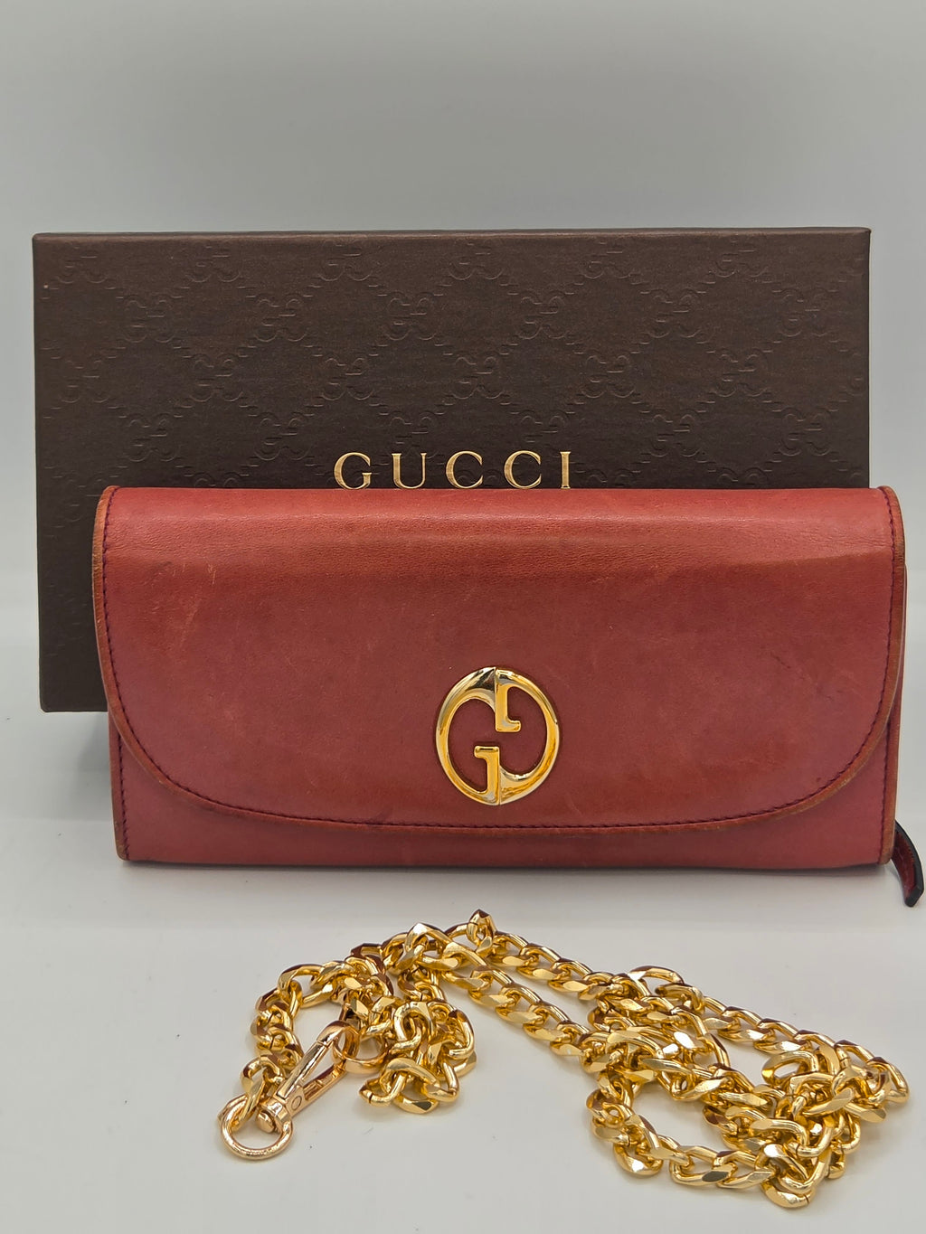A brown leather Gucci wallet with a gold-tone interlocking G logo, shown next to its dark brown Gucci box and a gold chain strap on a light gray background.