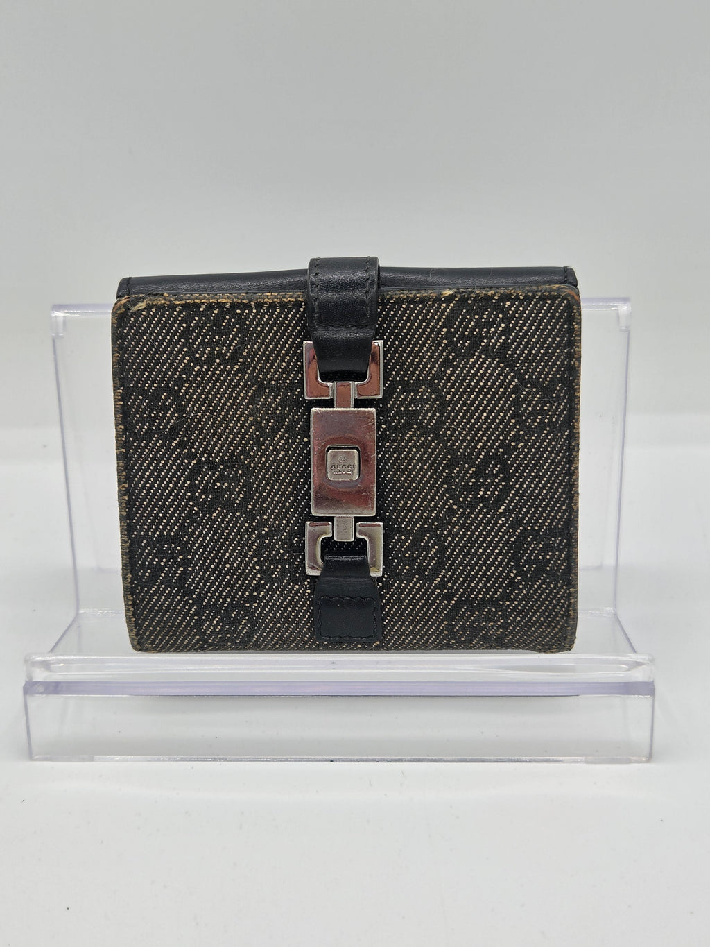 A dark brown and black monogram canvas bi-fold wallet with a black leather strap and silver-tone metal clasp, mounted on a clear acrylic display stand against a white background.