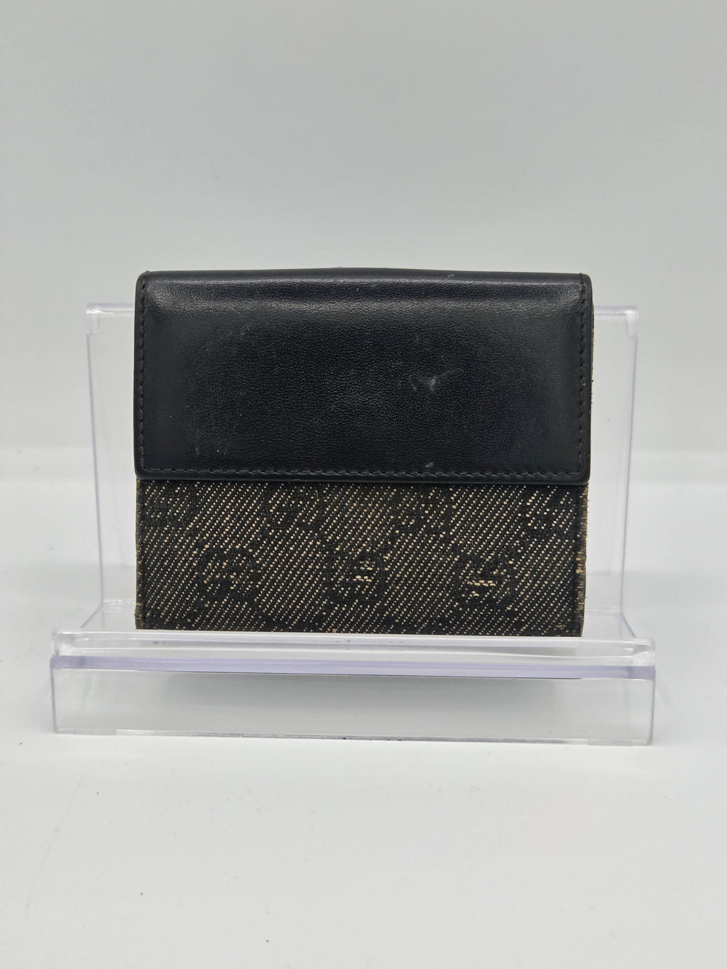 A black leather and monogram canvas bi-fold wallet displayed in a clear acrylic stand against a plain white background.